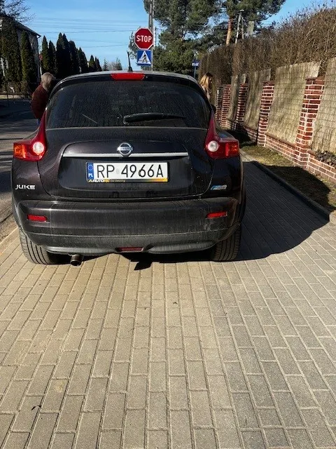Czarny samochód Nissan Juke z tyłu, z widoczną tablicą rejestracyjną RP 4966A. W tle znak STOP, niebieski znak drogowy z białym symbolem, ceglany mur, drzewa i domy. Samochód stoi na brukowanej powierzchni, niebo jest bezchmurne.