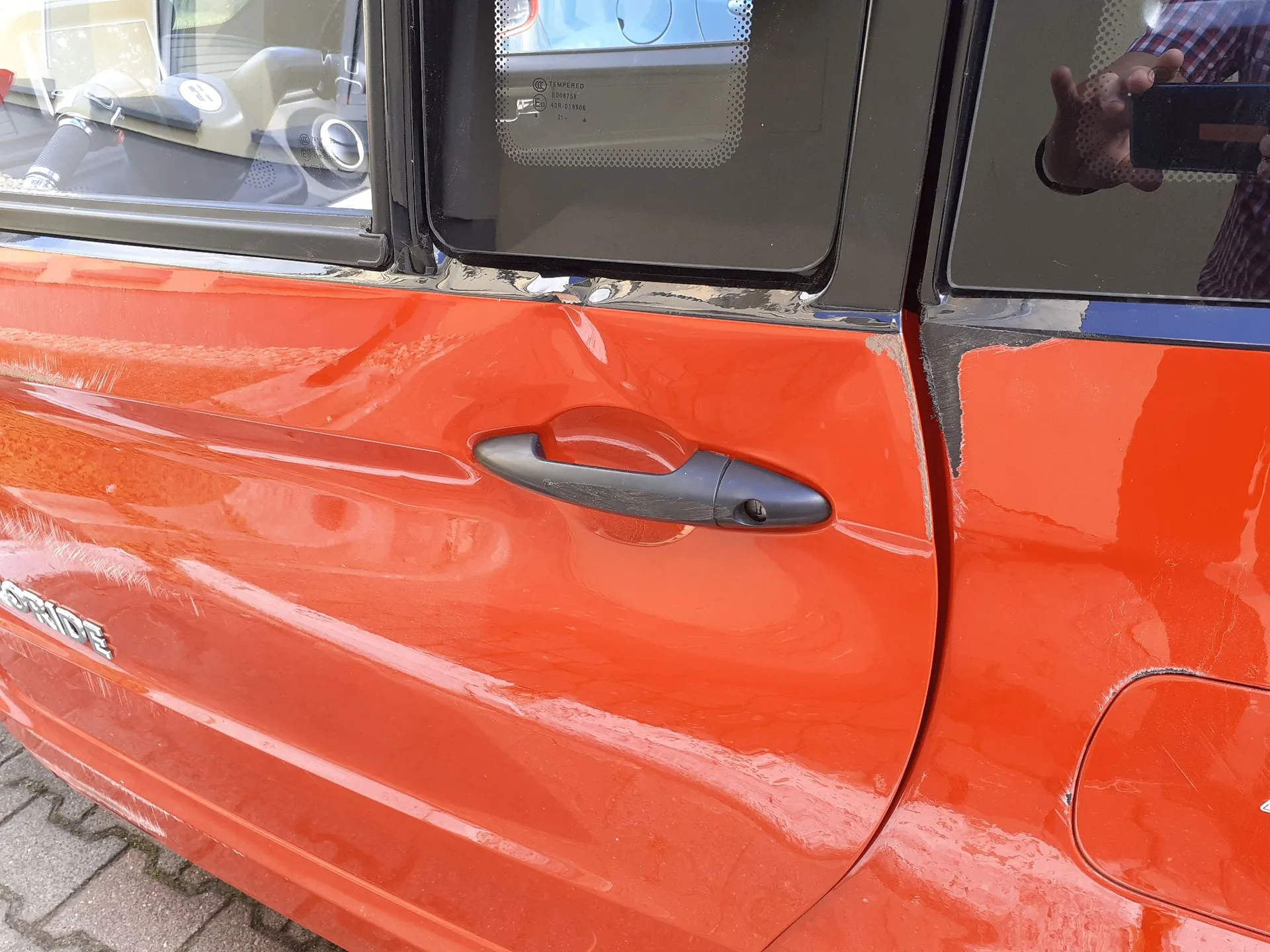 Zbliżenie na pomarańczowe drzwi samochodu z widocznym wgnieceniem w okolicach klamki. Klamka jest czarna i plastikowa. Powierzchnia lakieru jest porysowana, a w górnej części, przy krawędzi szyby, widoczne są odkształcenia blachy. W górnej części obrazu widoczna jest szyba z napisem 'TEMPERED'.