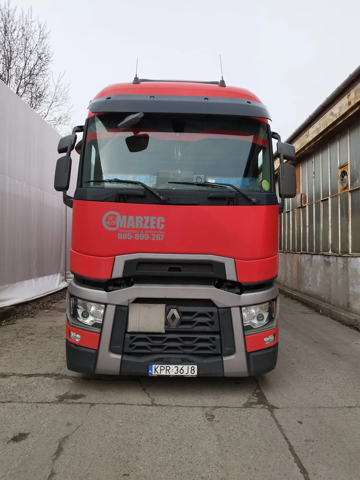 Przedni widok czerwonego ciężarówki marki Renault. Na masce widoczny napis "MARZEC TRANSPORT & SPEDITION 605-899-267". Tablica rejestracyjna KPR 36 J8. W szybie przedniej naklejki z napisami "DELTA SPED" po lewej stronie, "GRZES" po prawej oraz zielona naklejka z cyfrą "6". Po lewej stronie biały płot, po prawej przemysłowy budynek z metalowymi oknami. Niebo zachmurzone, podłoga z betonu.