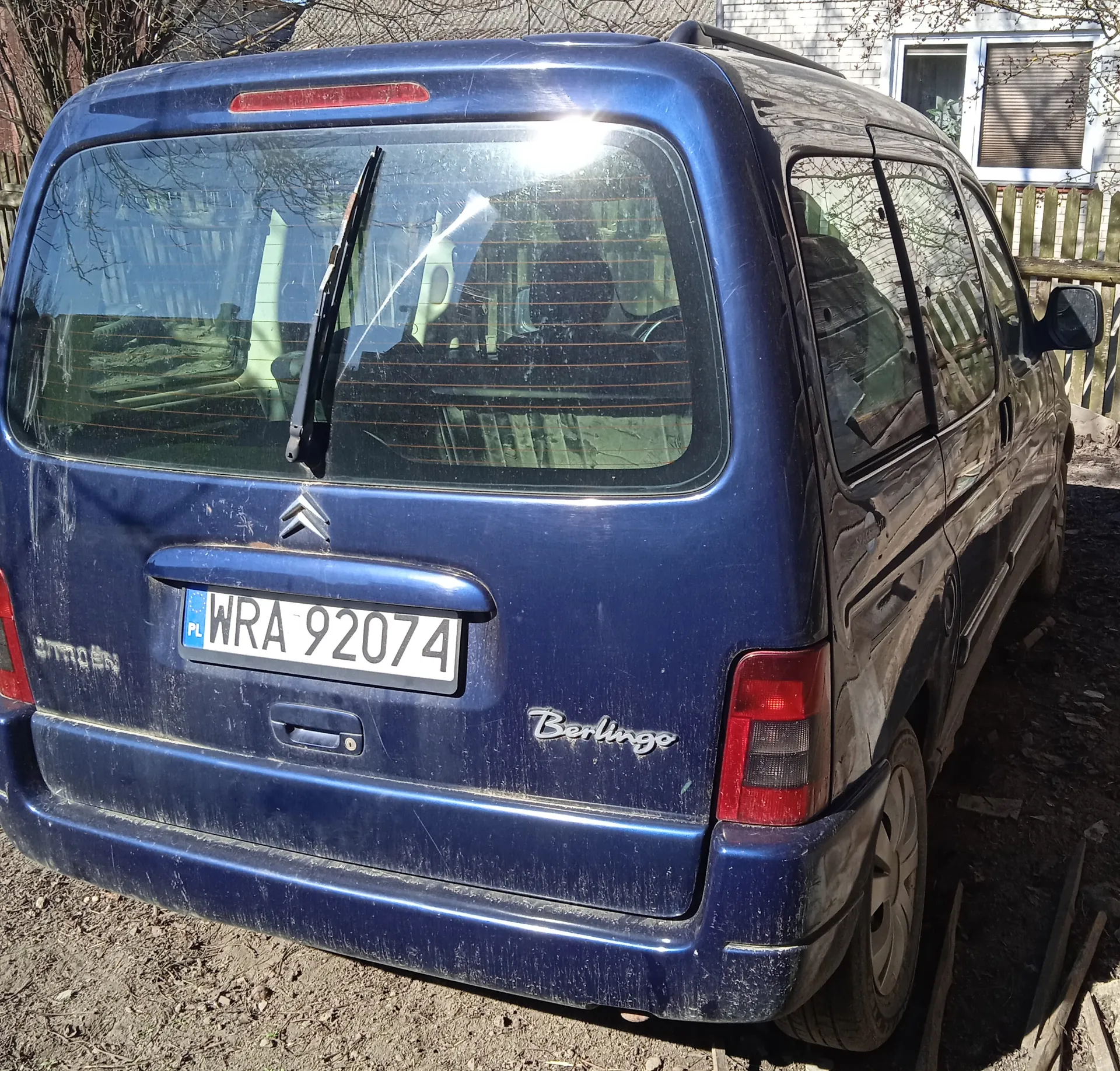 Niebieski samochód Citroën Berlingo z tyłu, z tablicą rejestracyjną WRA 92074 (PL). Widoczne są czerwone światła, logo marki, napis Berlingo oraz brudna karoseria. W tle dom z białą cegłą, drewniany płot i suche drzewa. Podłoga ziemna z suchą trawą.