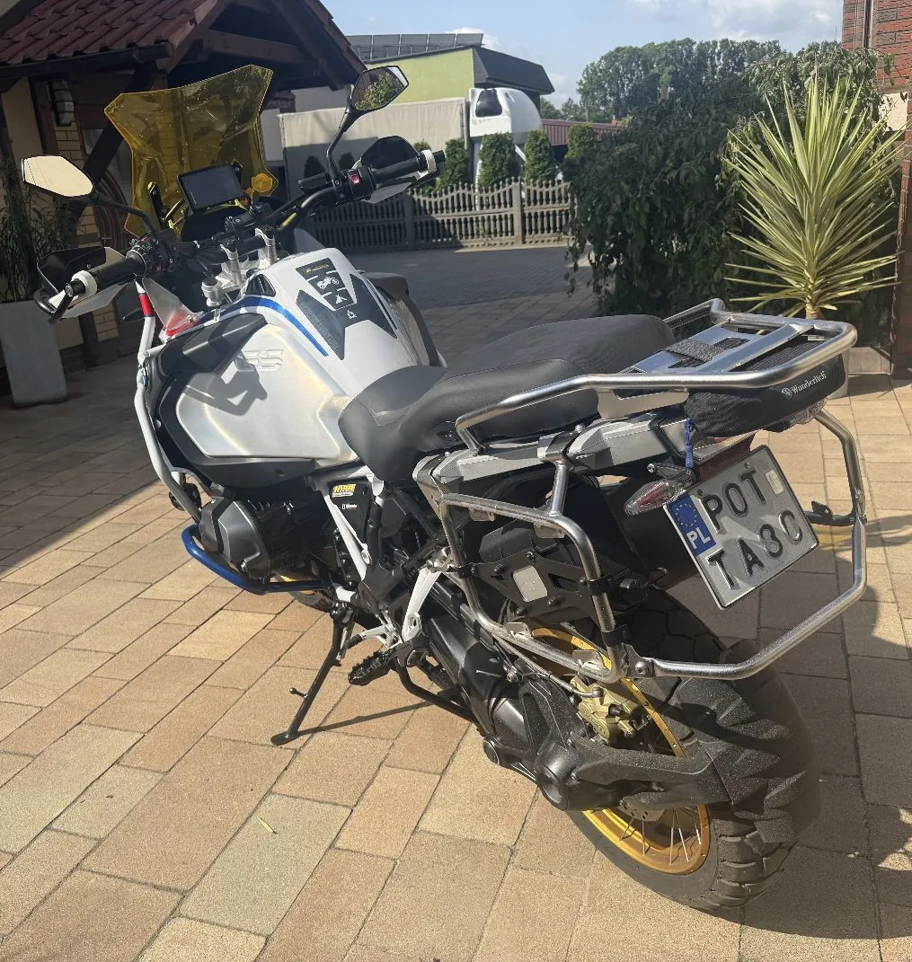 Srebrny motocykl turystyczny zaparkowany na kostce brukowej przed domem. Motocykl posiada czarną kanapę, srebrny bagażnik z tyłu oraz tablicę rejestracyjną o numerze POT TA30. W tle widoczny jest fragment budynku, drewniany płot oraz zielone rośliny, w tym palma.