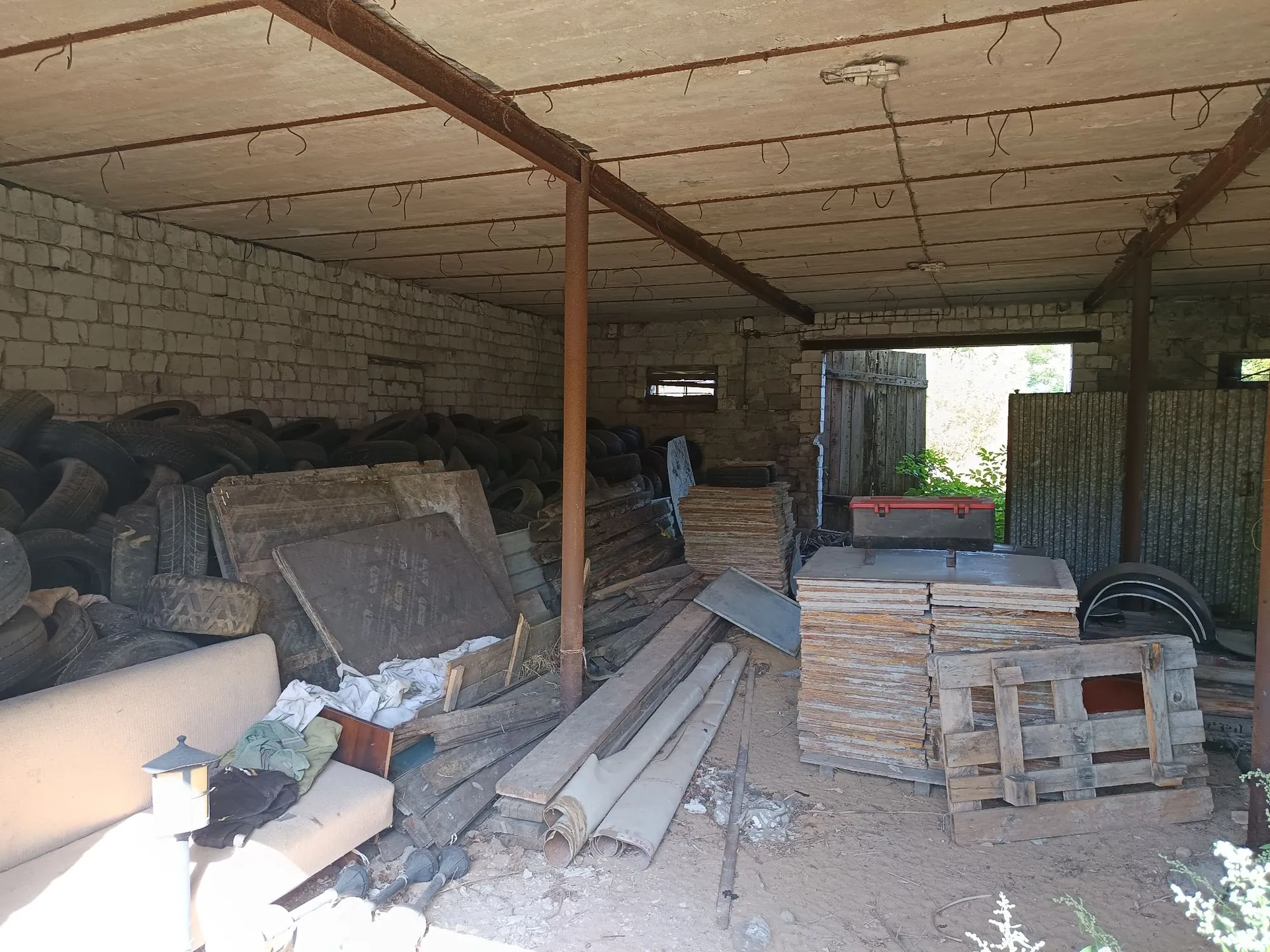 Wnętrze starego, zakurzonego garażu lub magazynu z betonowym sufitem wspartym na metalowych belkach. Po lewej stronie znajduje się duża sterta zużytych opon samochodowych. W centrum i po prawej stronie widoczne są stosy drewnianych płyt, desek, palet oraz metalowych elementów konstrukcyjnych. W lewym dolnym rogu stoi fragment białej kanapy z rzuconymi na niej szmatami. W tle widać otwarte wejście prowadzące na zewnątrz, przez które wpada światło dzienne.