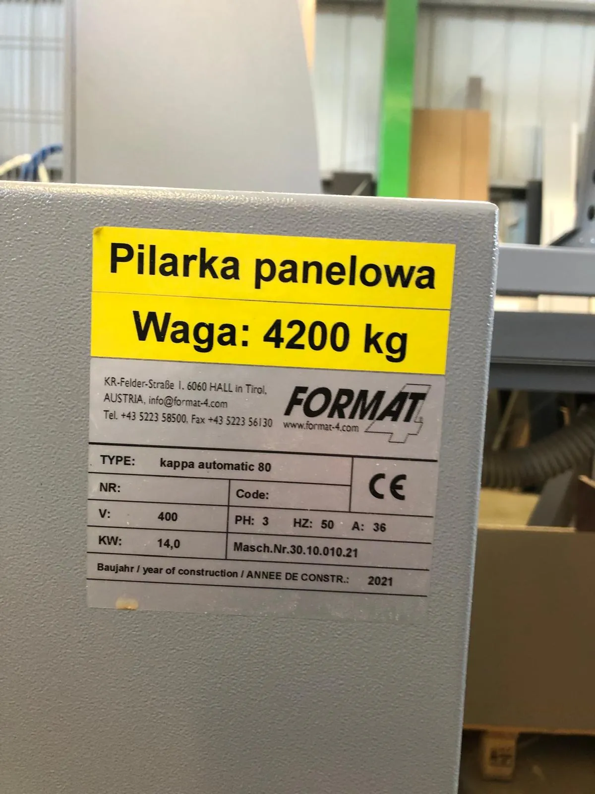 Zbliżenie na tabliczkę znamionową przymocowaną do szarej maszyny przemysłowej. Na górze znajduje się żółta naklejka z napisem: 'Pilarka panelowa Waga: 4200 kg'. Poniżej znajduje się srebrna tabliczka z danymi technicznymi: producent 'FORMAT', adres w Austrii, e-mail info@format-4.com, numer telefonu +43 5223 58500, faks +43 5223 561/30 oraz strona www.format-4.com. Specyfikacja techniczna obejmuje: TYPE: kappa automatic 80, V: 400, PH: 3, HZ: 50, A: 36, KW: 14,0, Masch.Nr. 30.10.010.21 oraz Baujahr / year of construction / ANNEE DE CONSTR.: 2021. Widoczne jest również oznaczenie CE.