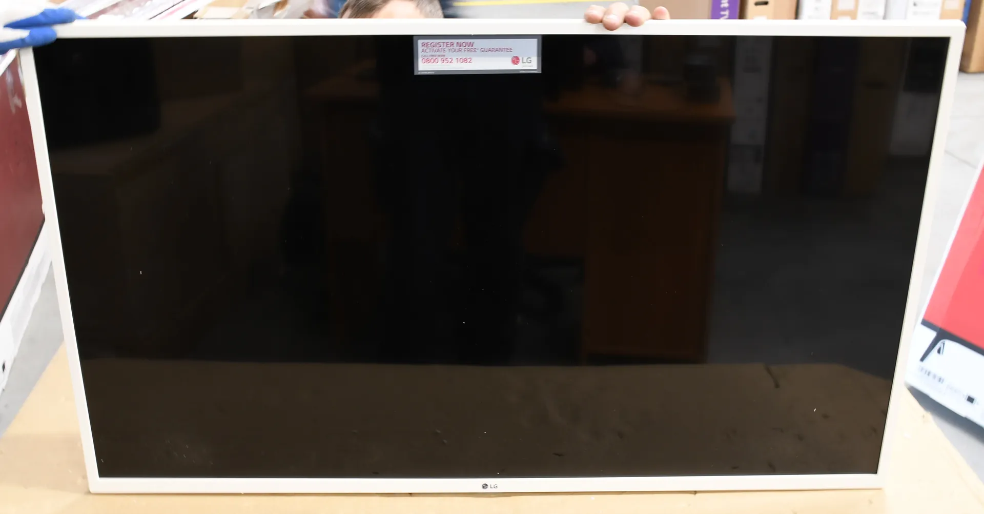 Zdjęcie przedstawiające z przodu biały monitor lub telewizor LG z czarnym ekranem. Na górnej krawędzi ramki znajduje się naklejka z napisem 'REGISTER NOW ACTIVATE YOUR FREE GUARANTEE' oraz numerem telefonu '0800 952 1082' i logo LG. Na dole ramki widoczne jest małe logo LG.