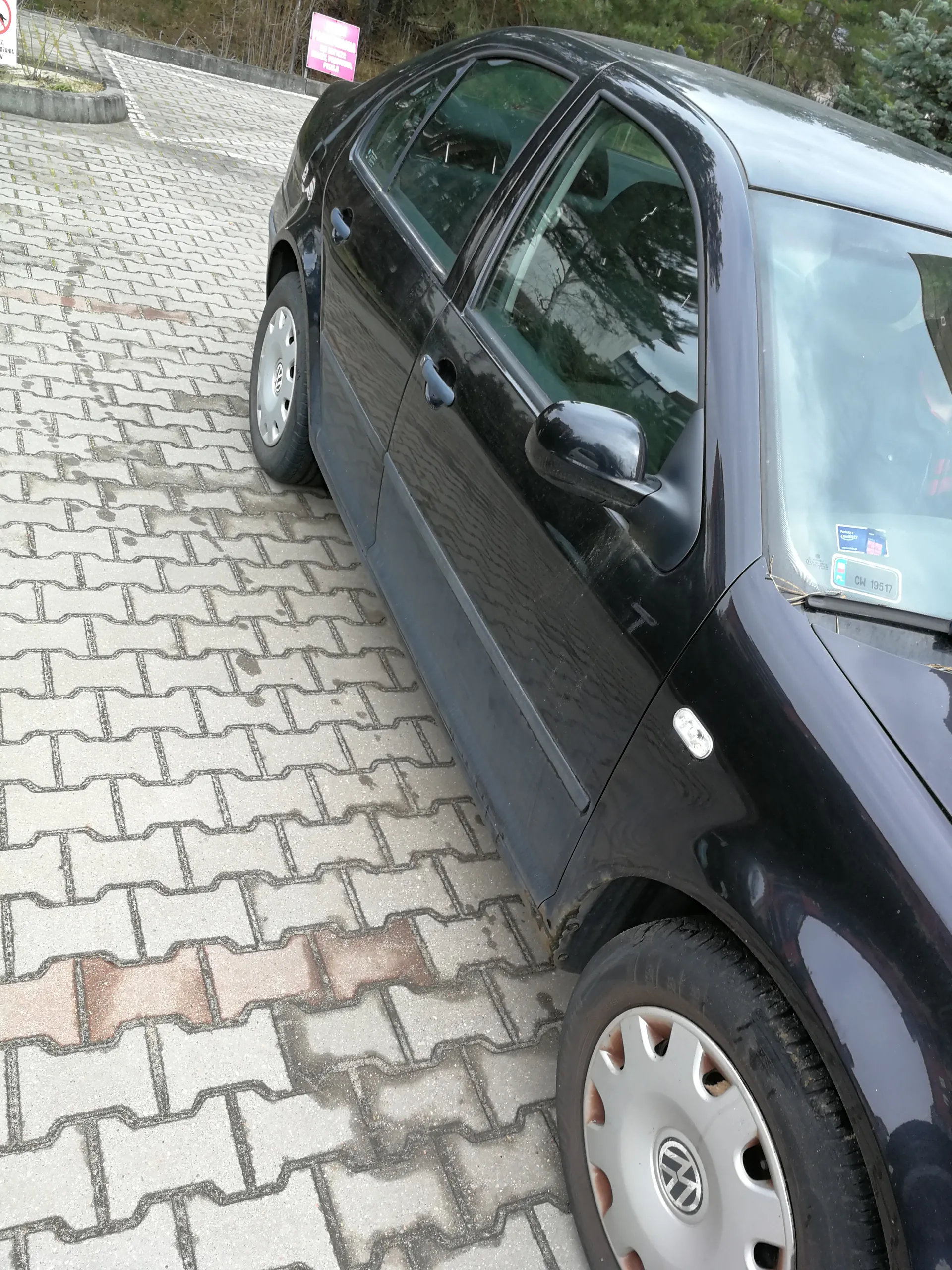 Czarny samochód osobowy marki Volkswagen zaparkowany na chodniku z kostki brukowej. Widoczna tablica rejestracyjna na przedniej szybie z napisem CM 19517 oraz niebieska naklejka. W tle różowy znak informacyjny i drzewa.