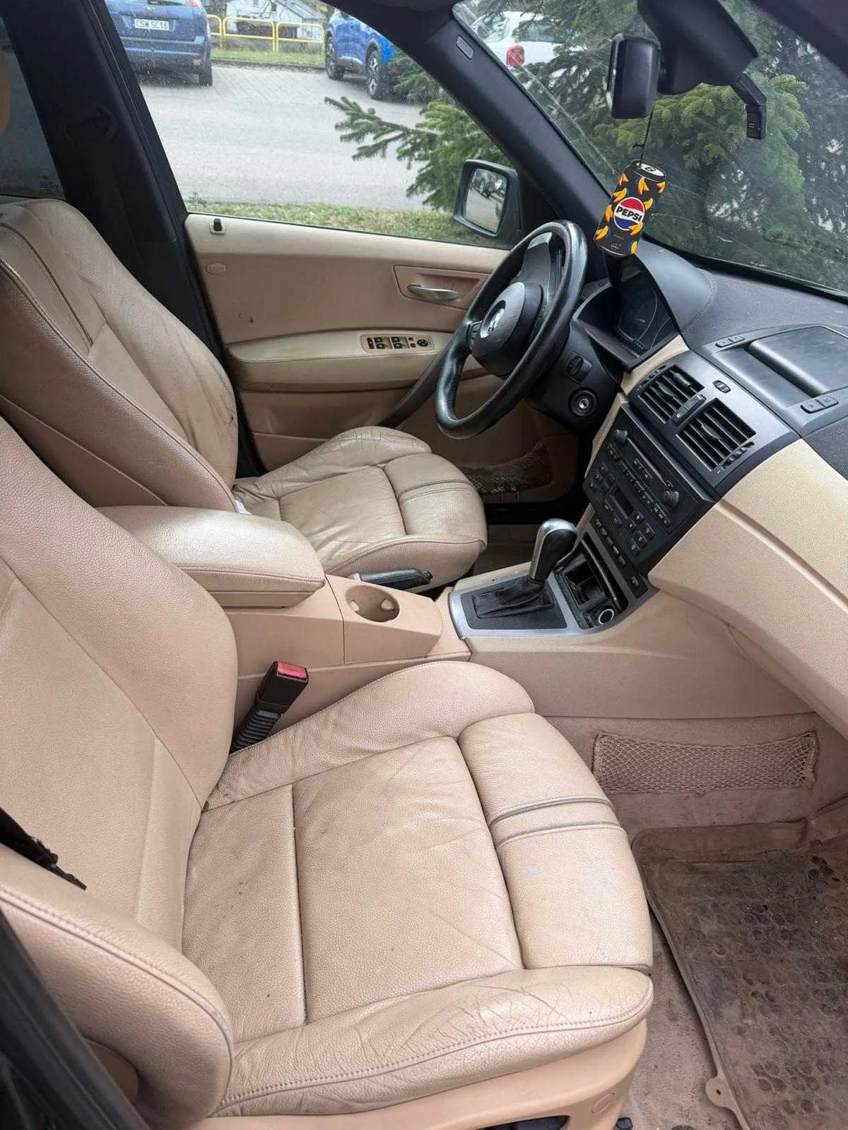 Wnętrze samochodu z beżowymi skórzany siedzeniami, kierownicą po lewej stronie, przekładnią biegów i deska rozdzielczą z czarnymi wentylacjami i przyciskami. Na lusterku wstecznym wiszący aromatyzator w kształcie puszki Pepsi z widocznym napisem "Pepsi". Przez przednią szybę widoczne zaparkowane samochody i drzewa. W tle fragment asfaltowej drogi i żółte barierki.