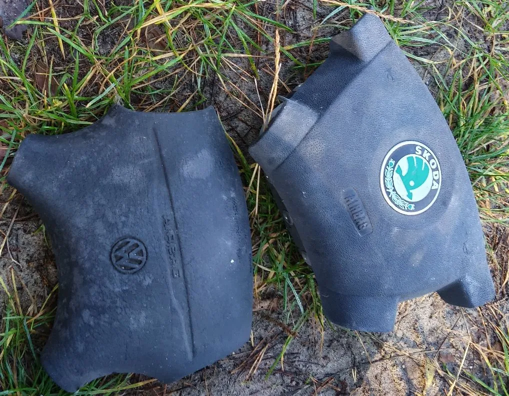 Dwie czarne, plastikowe osłony poduszek powietrznych z kierownic samochodowych, leżące na trawie. Lewa osłona posiada logo Volkswagena oraz napis 'AIRBAG'. Prawa osłona posiada logo marki Skoda.