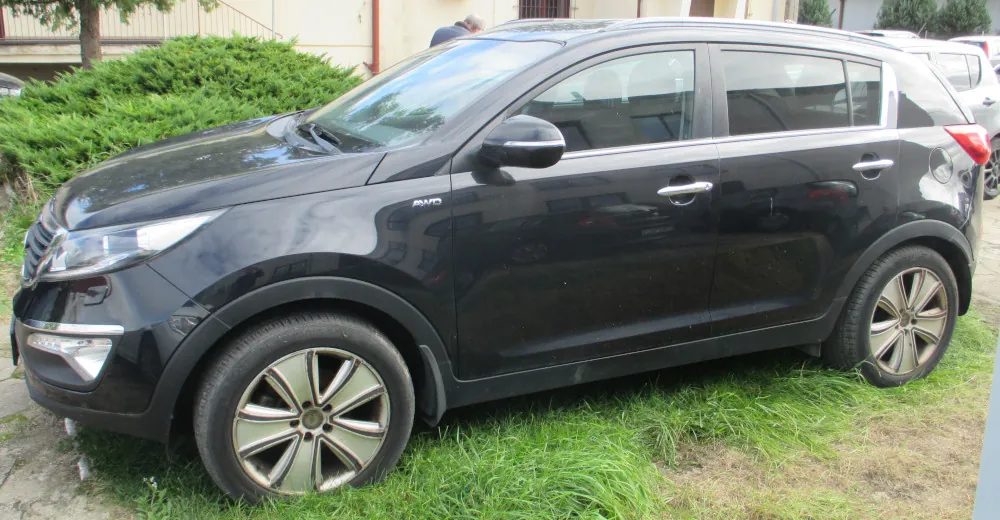 Czarny samochód typu SUV zaparkowany na trawie. Pojazd jest widoczny z profilu od strony kierowcy. Na przednim błotniku znajduje się mały napis 'AWD'. W tle widać zielone krzewy oraz fragment budynku.