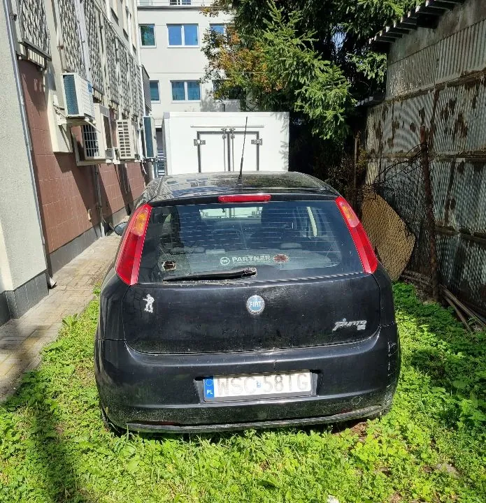 Tylny widok czarnego samochodu marki Fiat Punto zaparkowanego na trawie obok budynku. Samochód jest brudny, a na tylnej szybie znajduje się naklejka z napisem 'PARTNER'. Tablica rejestracyjna samochodu to WSC 58TG. W tle widoczny jest jasny budynek z jednostkami klimatyzacji na ścianie oraz zielone drzewa i metalowy płot.