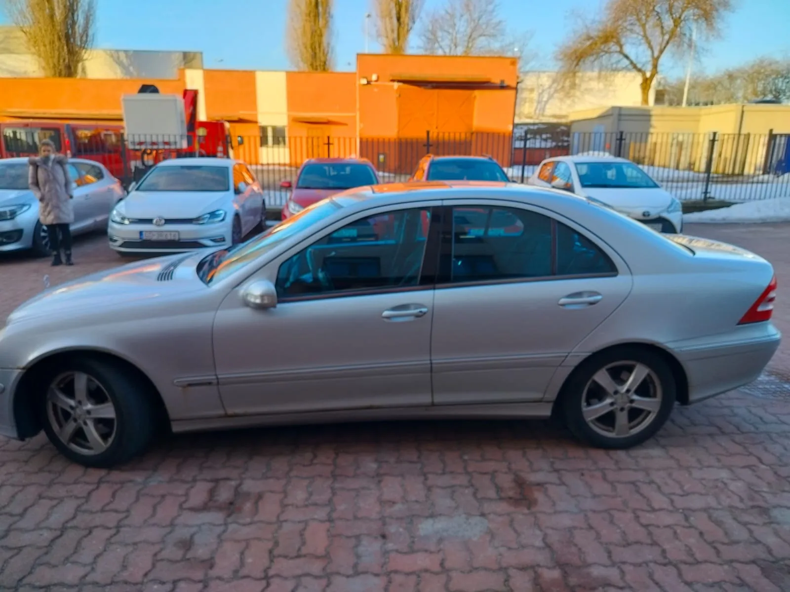 Srebrny samochód sedan zaparkowany na brukowanej powierzchni. W tle widoczne są inne pojazdy: biały Volkswagen, czerwony samochód oraz kolejny biały samochód. Po lewej stronie stoi osoba w długim płaszczu. Za pojazdami znajdują się pomarańczowe i beżowe budynki, metalowe ogrodzenie, drzewa bez liści oraz jasne niebo.