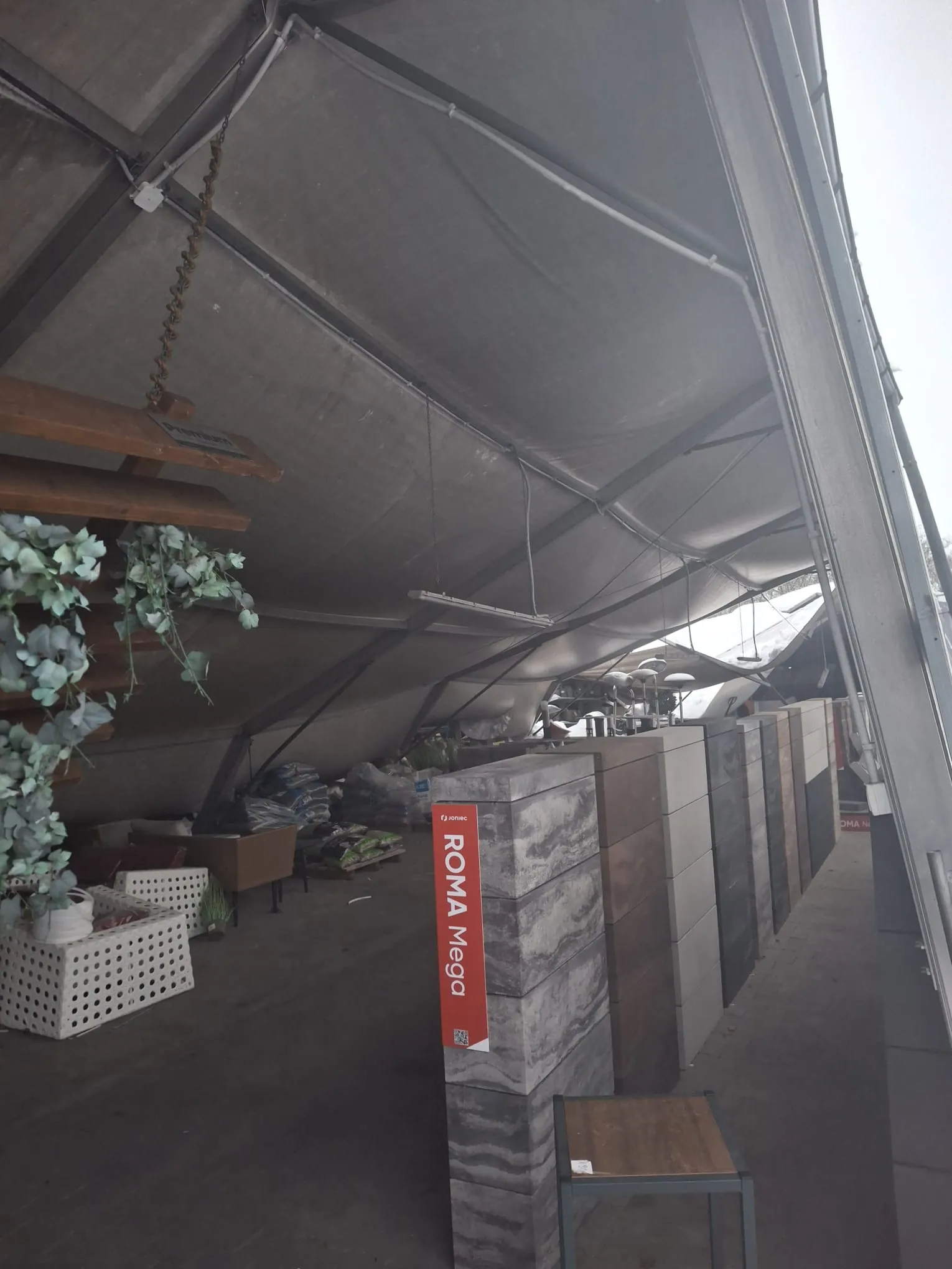 Wnętrze pomieszczenia pod dużym namiotem lub daszkiem. Po lewej stronie drewniane półki z wiszącymi zielonymi roślinami, poniżej białe kratowane pudełka, jedno z białym garnkiem. W centrum czerwony pionowy znak z napisem 'F. Jonec' oraz 'ROMA Mega', obok wysokie bloki z różnymi wzorami kamienia lub płytek. W tle stosy worków i przedmiotów, a także metalowe konstrukcje. Podłoga wyłożona płytkami, metalowe belki i szare tkaniny daszka.