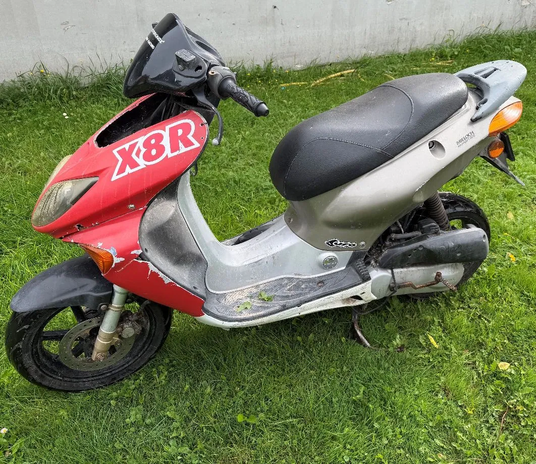 Srebrno-czerwony skuter marki X8R zaparkowany na zielonej trawie. Przednia owiewka jest czerwona z białym napisem 'X8R', a reszta nadwozia i siedzenie są w kolorze srebrnym i czarnym. Skuter jest widoczny z profilu, z lewej strony.