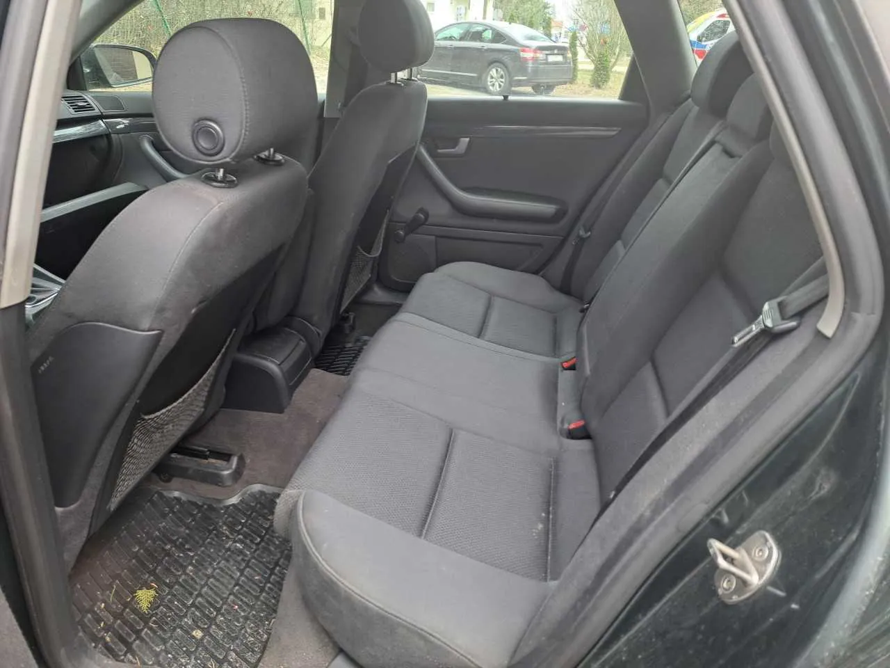 Wnętrze samochodu z widokiem na tylne siedzenia. Szare tkaninowe siedzenia z zagłówkami, pasy bezpieczeństwa i dywaniki podłogowe. Widoczne są przednie siedzenia, drzwi z uchwytem i okno. Na zewnątrz przez otwarte drzwi widać inny samochód oraz drzewa i krzewy na tle budynku.