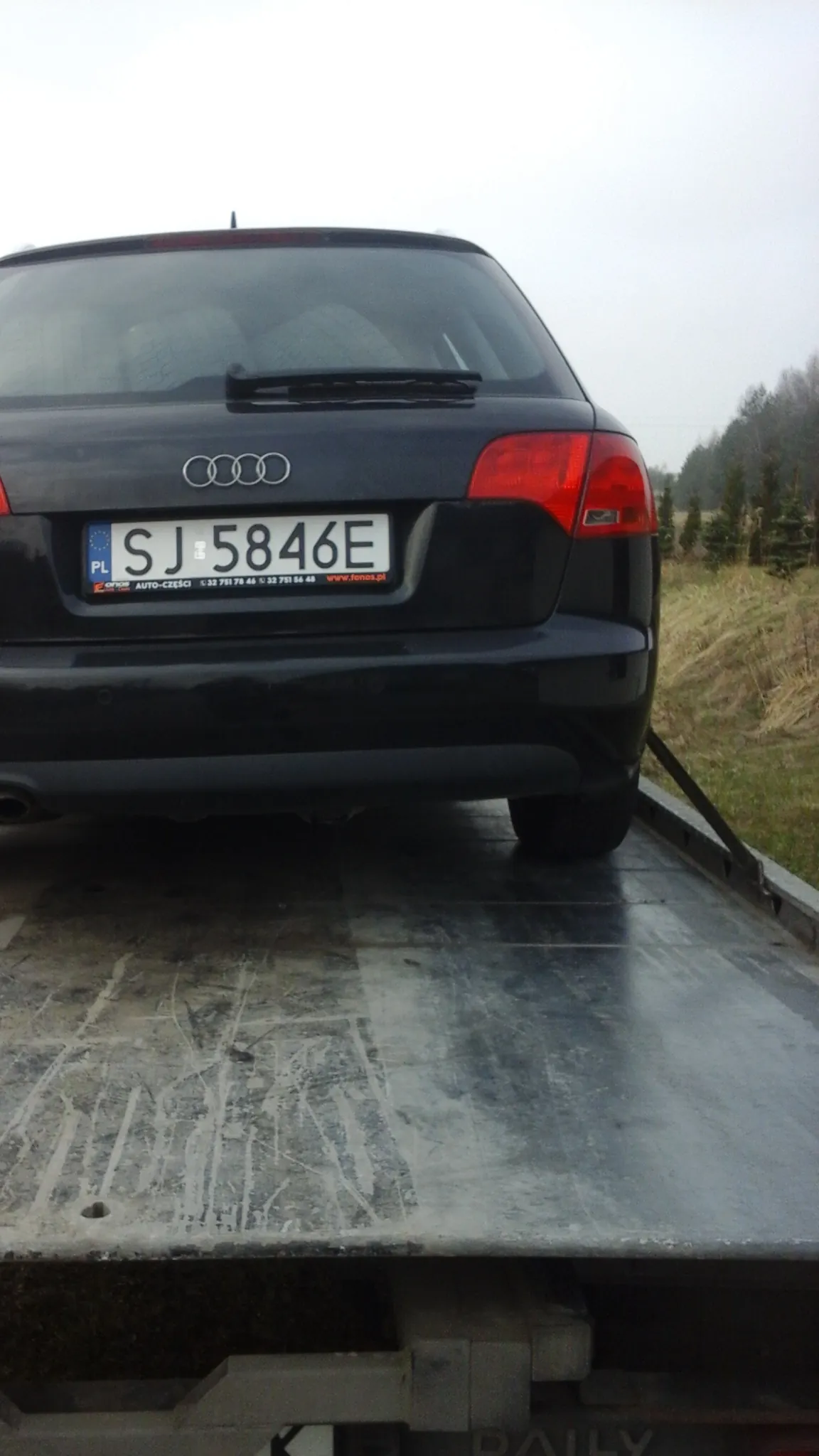 Czarny samochód marki Audi z tablicą rejestracyjną SJ 5846E na drodze. W tle trawa i drzewa. Tablica rejestracyjna zawiera oznaczenie 'PL' oraz dodatkowe napisy. Widoczne są czerwone światła tylnego reflektora i logo Audi z czterema pierścieniami.