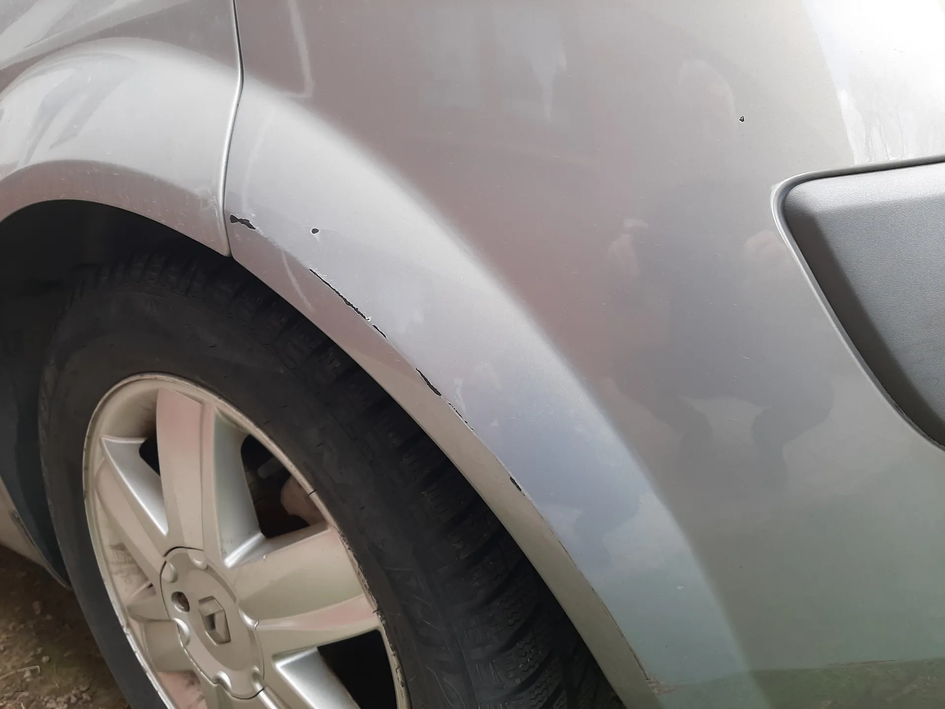 Zbliżenie na przednią prawą część srebrnego samochodu. Widoczne są uszkodzenia na błotniku w postaci zadrapań i małego wgniecenia. Część koła z oponą i aluminiową felgą. Na karoserii odbicie osoby robiącej zdjęcie. Brak czytelnego tekstu.