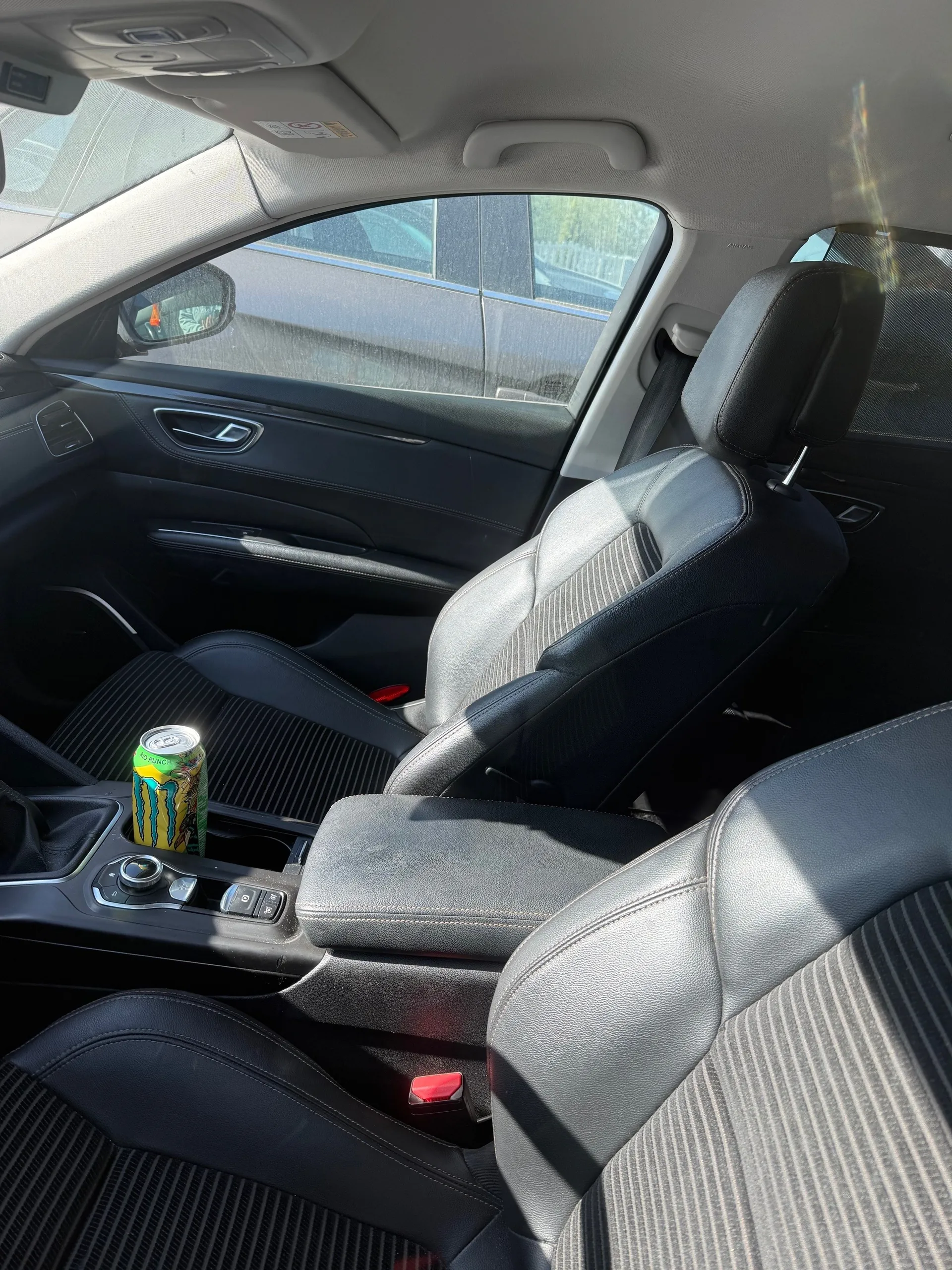 Wnętrze samochodu z widocznymi czarno-szarymi fotelami tapicerowanymi skórą w paski. W uchwycie na kubki w centralnej konsoli znajduje się kolorowa puszka z żółtym, zielonym i niebieskim wzorem. Widoczne są czarne panele drzwi z srebrnymi akcentami, lusterko boczne oraz fragment zewnętrzny z innym pojazdem i budynkiem za oknem. Słoneczne światło wpada przez szyby, tworząc refleksy.