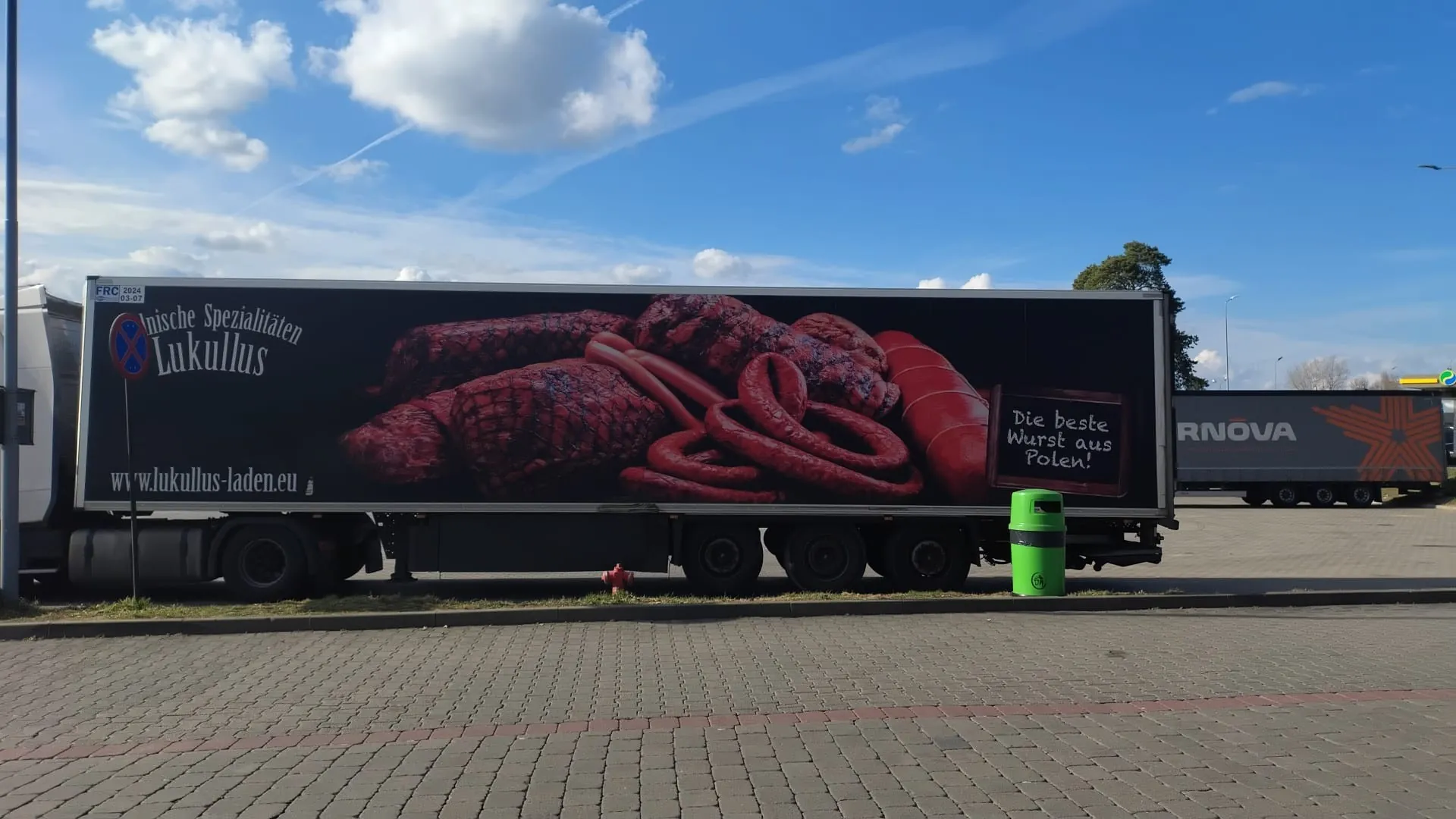Duży naczepa ciężarowa z reklamą przedstawiającą różne rodzaje kiełbas i wędlin na czarnym tle. Na lewej stronie napisy: 'Polnische Spezialitäten Lukullus' oraz 'www.lukullus-laden.eu'. Po prawej stronie tabliczka z napisem 'Die beste Wurst aus Polen!'. W tle niebo z białymi chmurami, druga naczepa z napisem 'RNOVA' i pomarańczową gwiazdą, zielony pojemnik na śmieci oraz brukowana powierzchnia. Widoczne są również drzewa i słup z niebieskim znakiem zakazu parkowania.