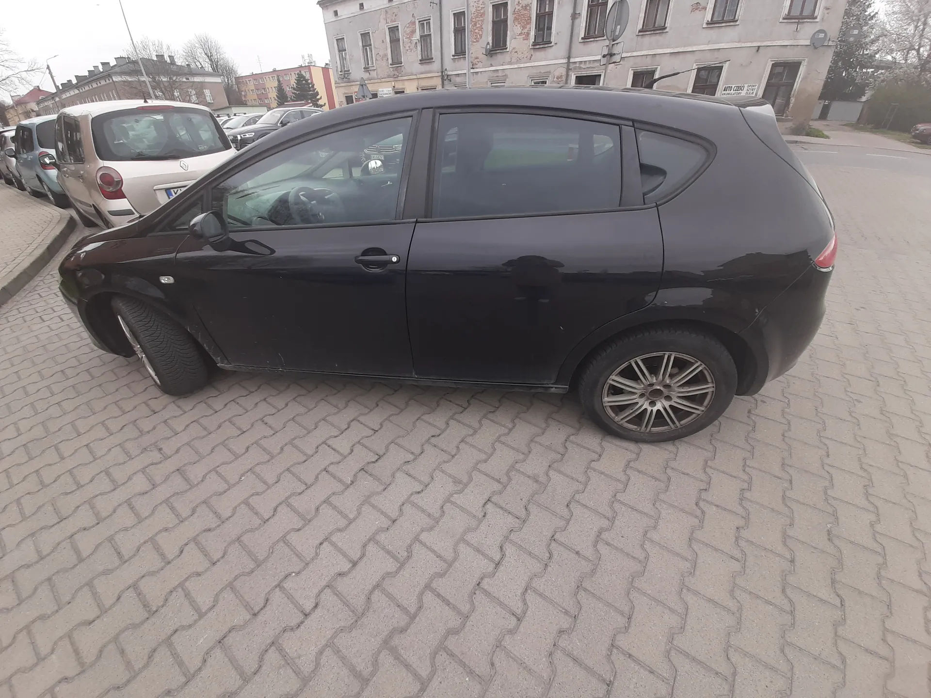 Czarny samochód typu hatchback zaparkowany na brukowanej powierzchni. W tle widoczne są inne zaparkowane pojazdy, w tym beżowy i niebieski samochód, oraz stary budynek z wieloma oknami i częściowo obłupioną elewacją. Na pierwszym planie brukowane tory o geometrycznym wzorze. Niebo jest pochmurne.