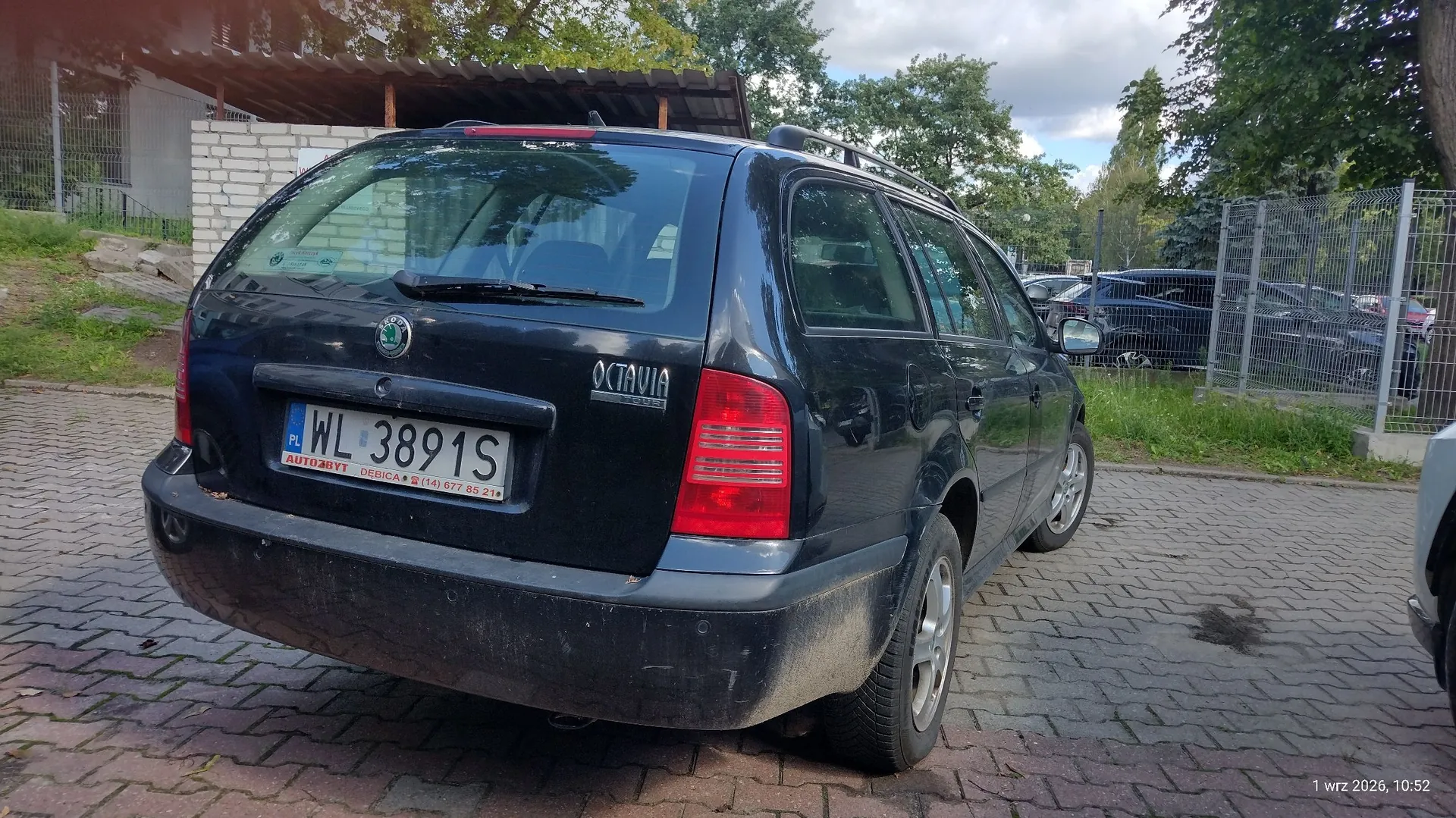 Tylny widok czarnego samochodu marki Skoda Octavia Combi zaparkowanego na kostce brukowej. Pojazd ma tablicę rejestracyjną o numerze WL 3891S. Na klapie bagażnika widoczne są emblematy marki Skoda oraz napis OCTAVIA. W tle widać ogrodzenie z siatki, drzewa oraz inny zaparkowany samochód.