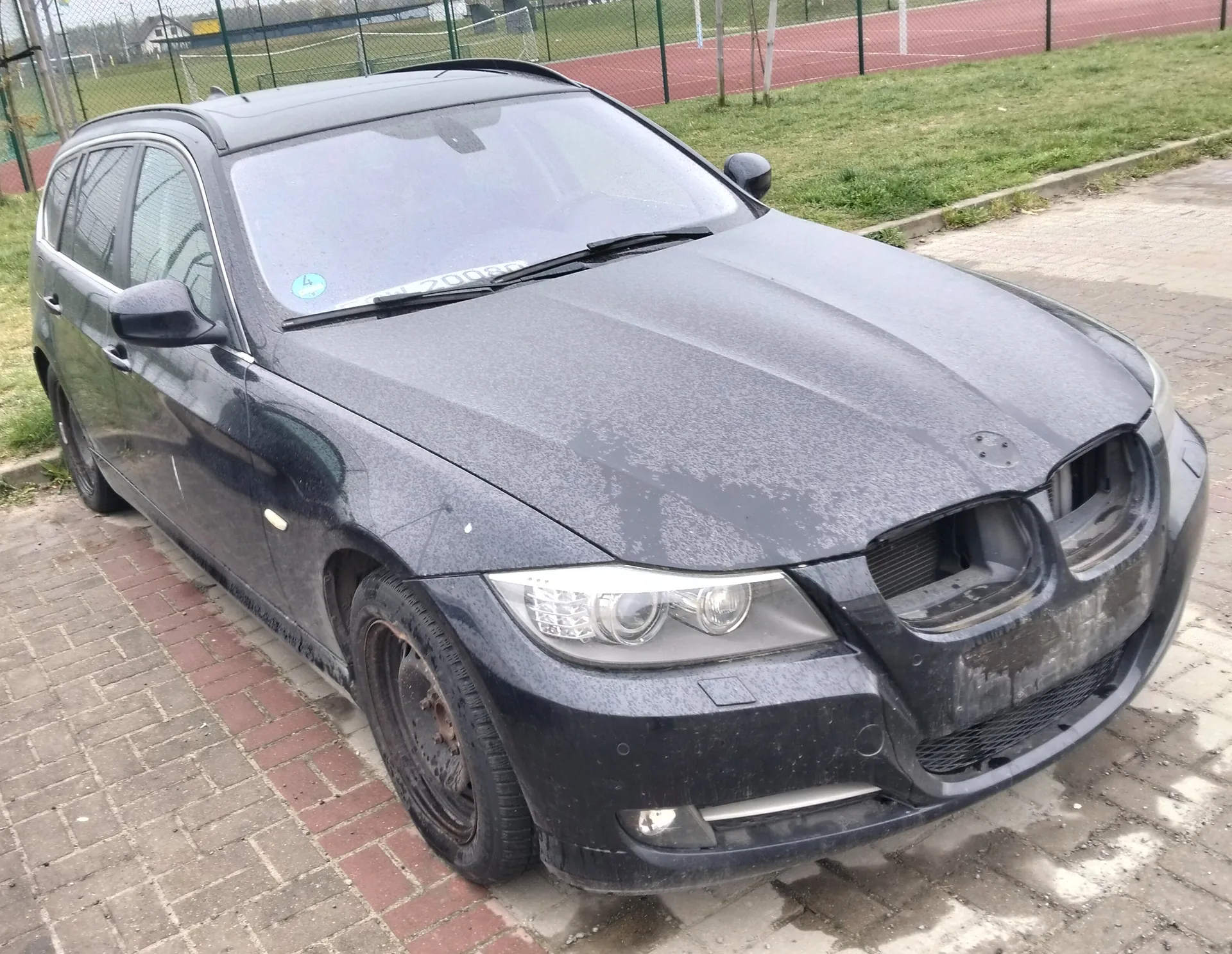 Czarny samochód typu kombi zaparkowany na brukowanej powierzchni. Pojazd jest zabrudzony, brakuje przedniego zderzaka i lewego przedniego koła. Na przedniej szybie widoczny niebieski naklejony znaczek. W tle boisko tenisowe z czerwoną nawierzchnią, zielone ogrodzenie, trawa oraz budynki w oddali.
