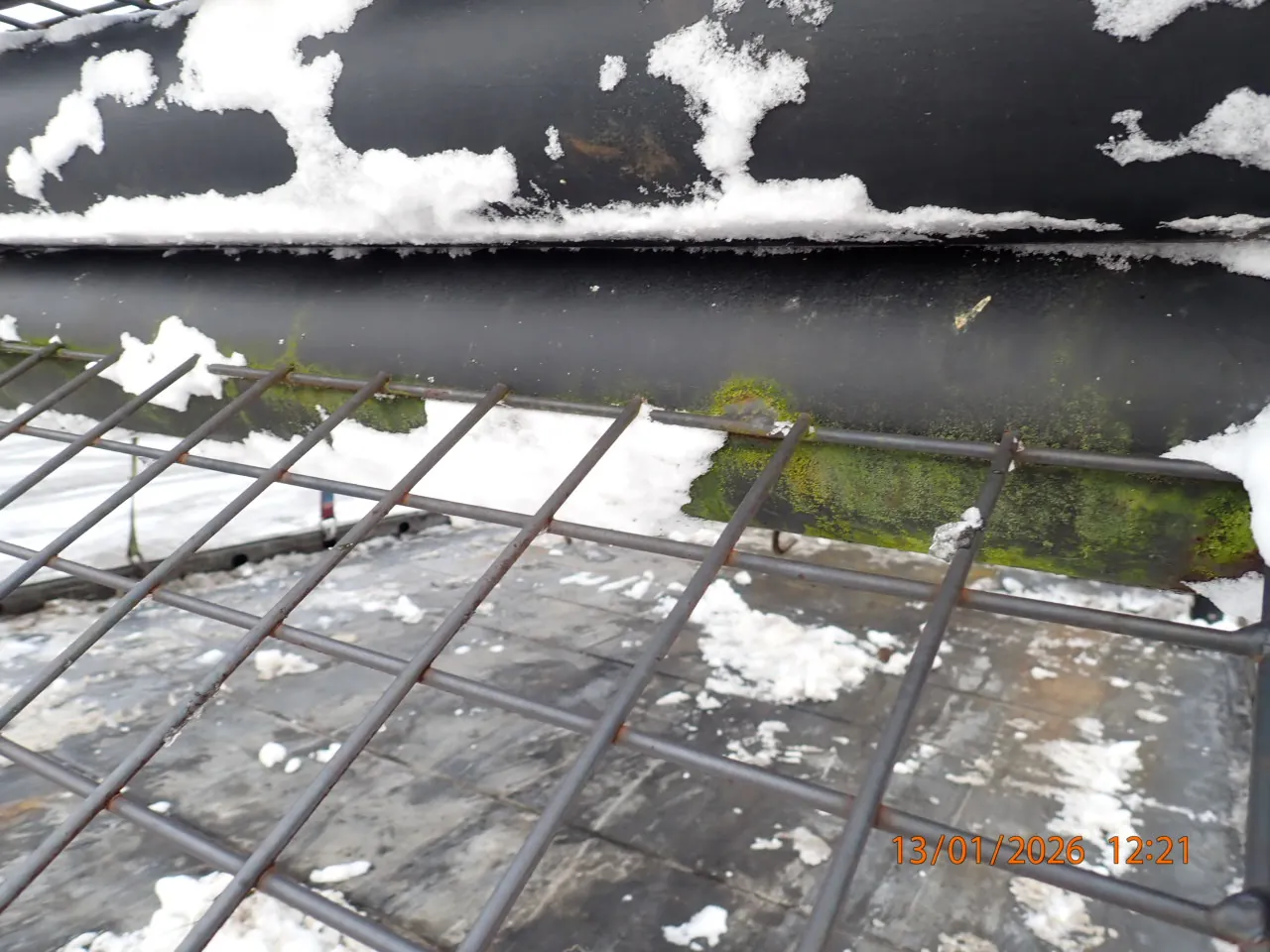 Czarny metalowy rurociąg pokryty śniegiem i zielonym nalotem, umieszczony nad metalową siatką. Na powierzchni poniżej siatki widoczne są resztki śniegu i brudna, wilgotna podłoga. W prawym dolnym rogu obrazu znajduje się pomarańczowy znaczek z datą i czasem: 13/01/2026 12:21.