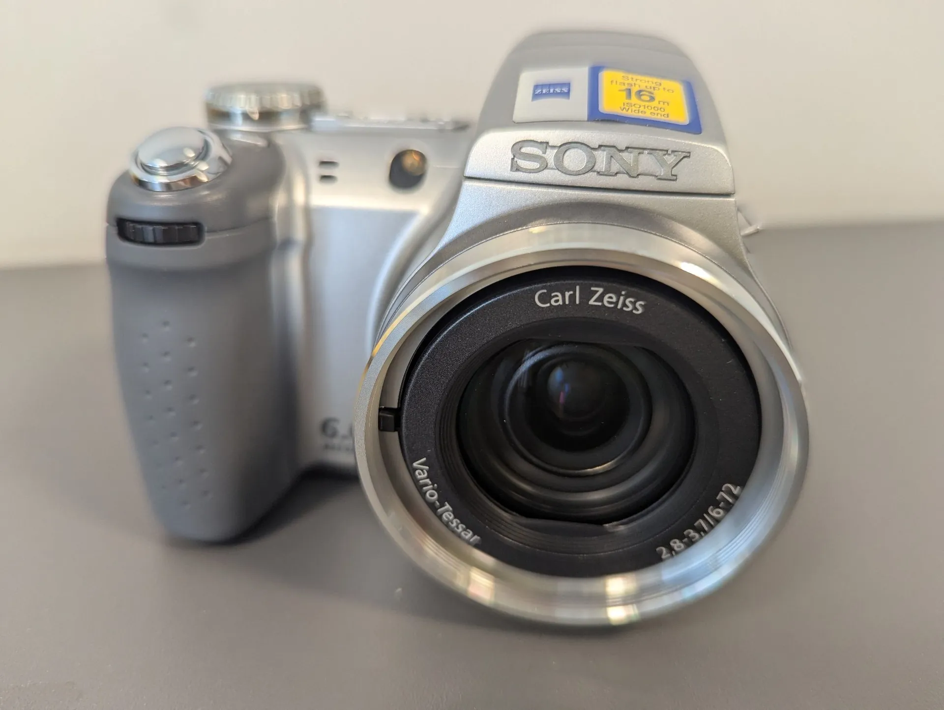 Srebrny aparat cyfrowy marki Sony z obiektywem Carl Zeiss Vario-Tessar. Na obudowie widoczne jest logo SONY oraz żółta naklejka z napisem 'Strong zoom 16x'. Obiektyw posiada oznaczenia 'Carl Zeiss', 'Vario-Tessar' oraz '2,8-3,7/6-72'.