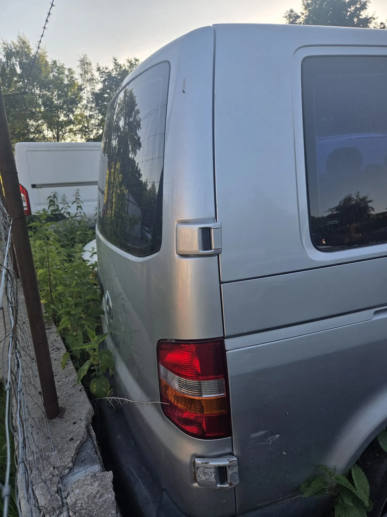 Srebrny van widziany od tyłu z lewej strony. Widoczne są czerwone i pomarańczowe światła tylnego, metalowa klamka na drzwiach oraz refleksy drzew w szybie. Po lewej stronie obrazu znajduje się siatka ogrodzeniowa z betonowymi blokami i zaroślami roślin. W tle biały van oraz drzewa na tle jasnego nieba.