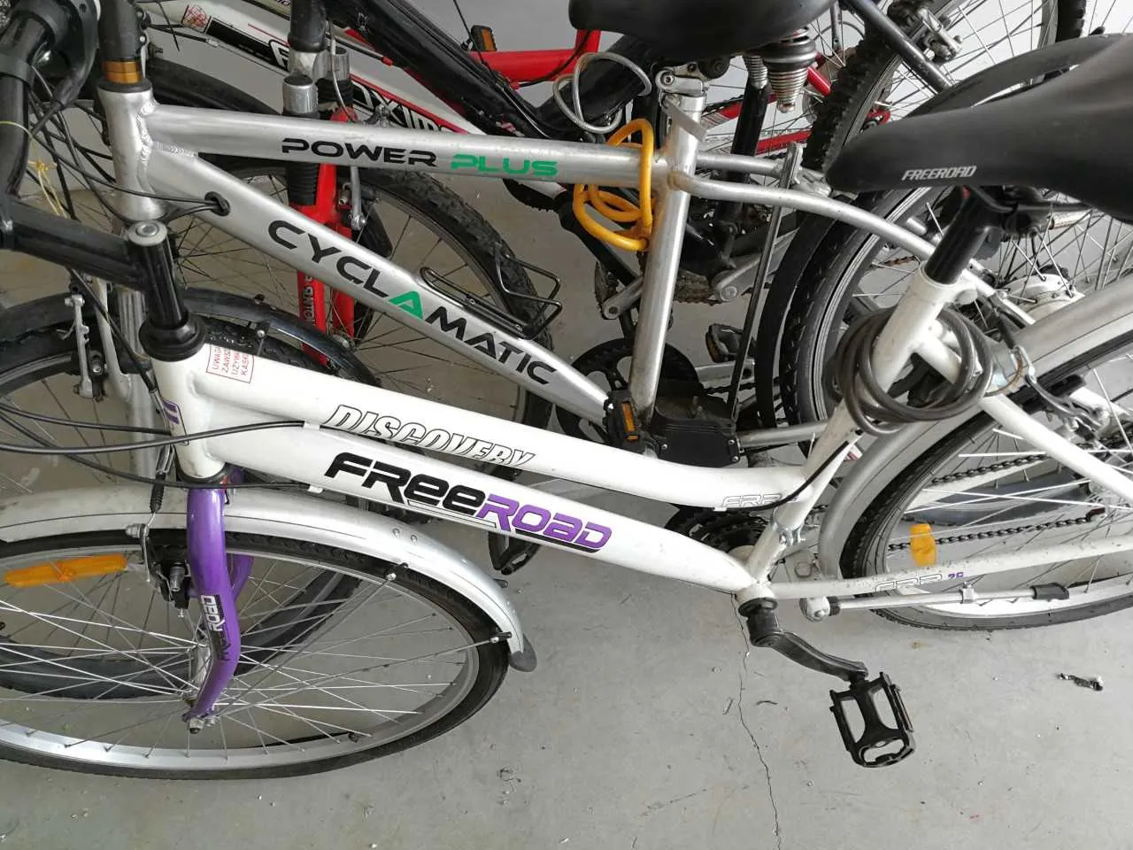 Zbliżenie na kilka rowerów zaparkowanych w pomieszczeniu z betonową podłogą. W pierwszym planie biały rower z napisami 'DISCOVERY' i 'FREEROAD' na ramie, fioletową widełką oraz czarnym siedziskiem. Za nim srebrny rower z napisami 'CYCLAMATIC' i 'POWER PLUS' (zielony napis 'PLUS'). Widoczne są również części innych rowerów, w tym czerwony rama i czarne koła. Na ramie białego roweru znajduje się pomarańczowy kabel zabezpieczający.