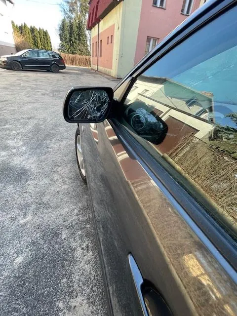 Zdjęcie przedstawia brązowy samochód z pękniętym lusterkiem bocznym, zaparkowany na podwórku z żwirem. W tle widoczny czarny samochód oraz różowo-żółty budynek z oknami. Na pierwszym planie lusterko boczne z widocznymi pęknięciami, a w odbiciu fragment budynku i suchego trawnika. Tło uzupełniają drzewa i jasne niebo.