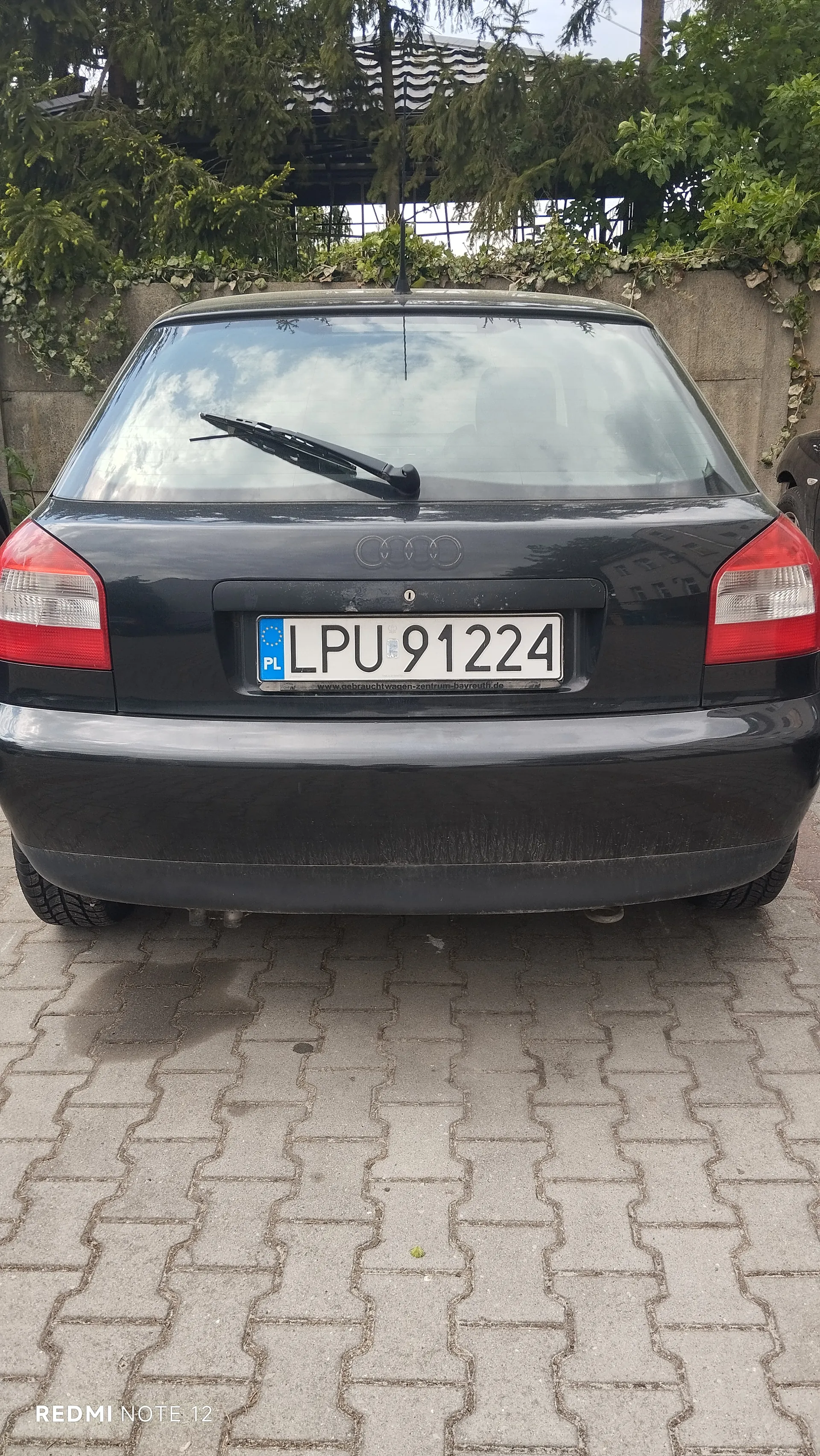 Tył czarnego samochodu marki Audi z tablicą rejestracyjną LPU 91224. Tablica posiada niebieski pas z napisem PL oraz białe tło z czarnymi literami i cyframi. Samochód stoi na brukowanej powierzchni, w tle widoczny kamienny mur porośnięty pnączami, drzewa i fragment budynku z dachem. W lewym dolnym rogu obrazu znajduje się napis REDMI NOTE 12.