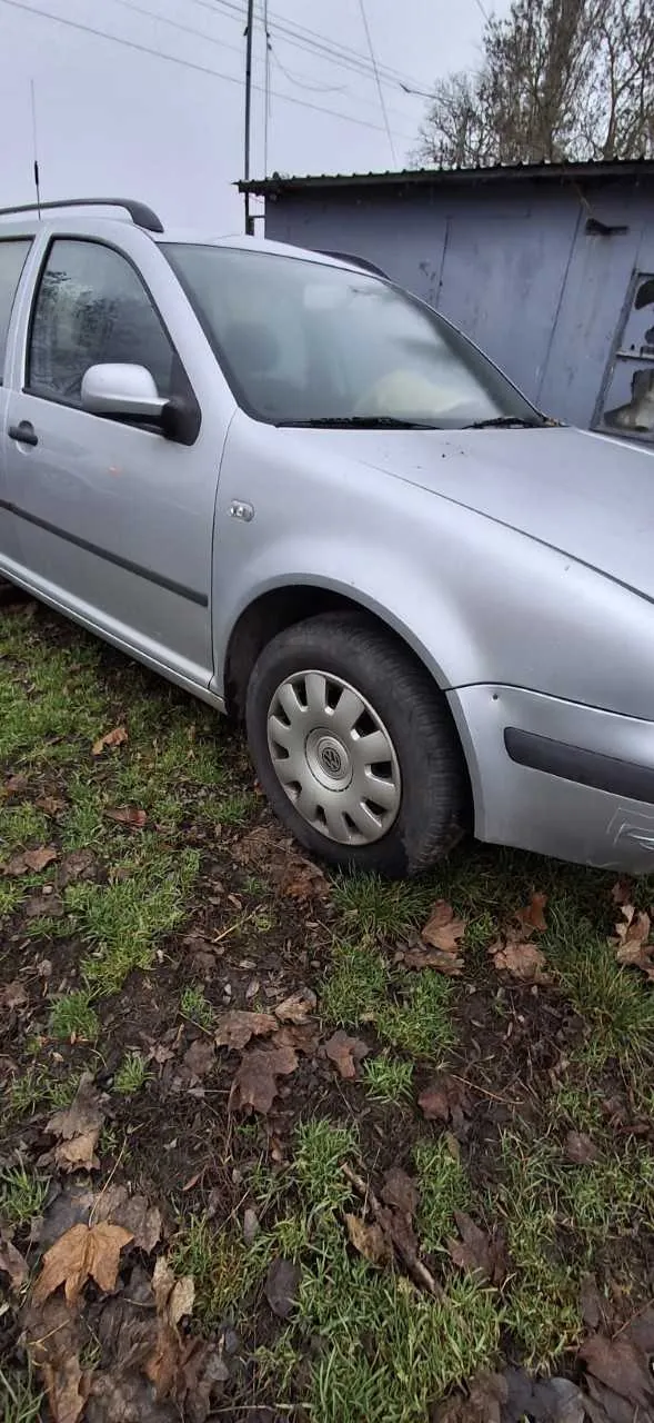 Srebrny samochód osobowy zaparkowany na trawie z suchymi liśćmi. W tle niebieski budynek gospodarczy, drzewa i słup z przewodami. Niebo jest zachmurzone.
