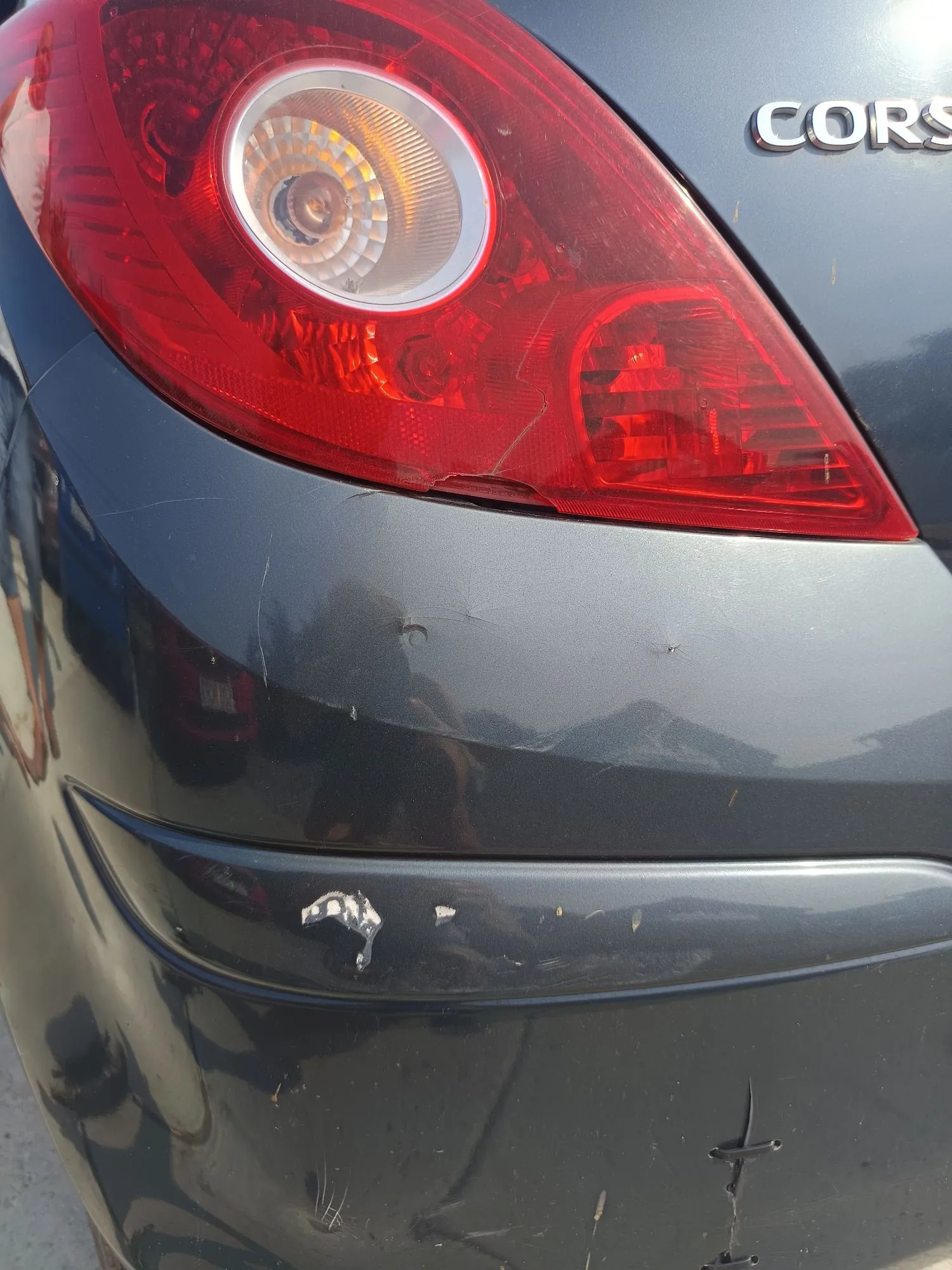 Zbliżenie na tylną lampę samochodu w kolorze ciemnoszarym. Lampa ma czerwone obudowy z białym, okrągłym elementem wewnątrz. Na karoserii powyżej lampy widoczny jest fragment napisu „CORS”. Na zderzaku poniżej lampy widoczne są liczne zarysowania i wgniecenia.
