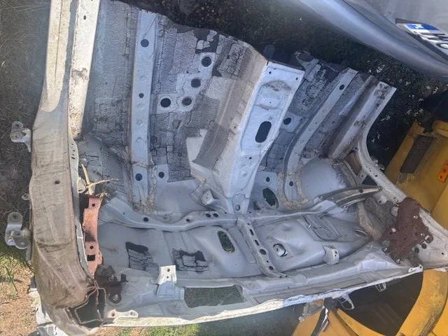 Zdjęcie przedstawia rozebrany metalowy element karoserii samochodu, prawdopodobnie drzwi lub przednią część, z widocznymi otworami, śladami korozji i uszkodzeniami. W tle częściowo widoczny żółty pojazd oraz trawa na ziemi. Na lewym górnym rogu widoczny fragment tablicy rejestracyjnej z niewyraźnymi literami i cyframi.
