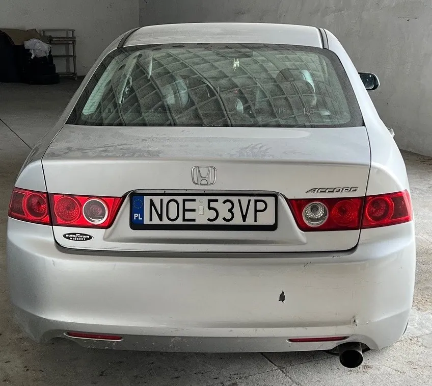 Tył srebrnego samochodu Honda Accord z tablicą rejestracyjną PL NOE 53VP. Widoczne są czerwone światła tylnego oświetlenia, logo Hondy, napis 'Accord' po prawej stronie bagażnika oraz niewielki czarny ślad na karoserii poniżej tablicy. Samochód zaparkowany w pomieszczeniu z betonowymi ścianami i podłogą. W tle widoczne są elementy wyposażenia garażowego.