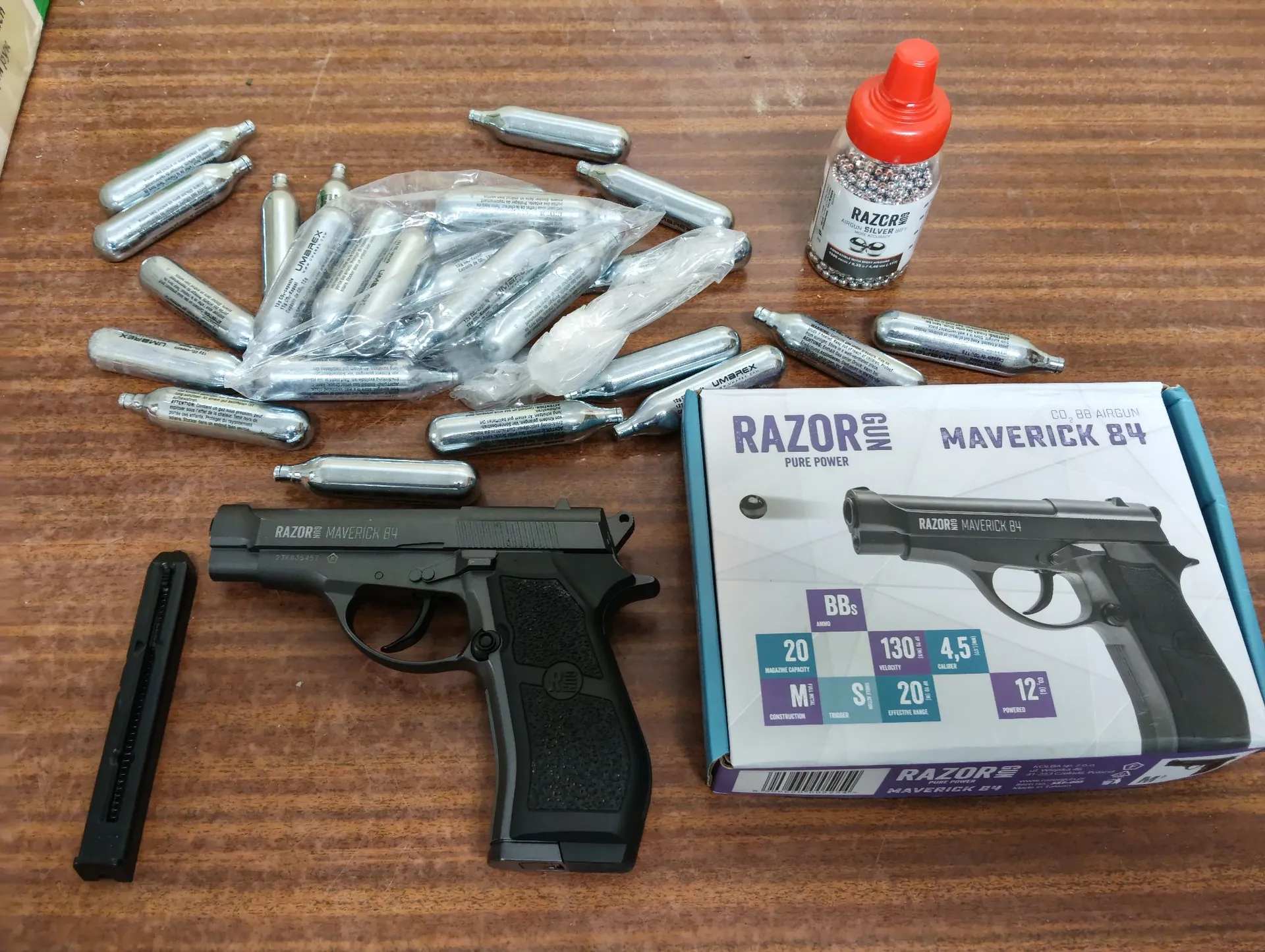 Na drewnianym blacie leży czarny pistolet pneumatyczny Razor Maverick B4, obok niego znajduje się jego oryginalne biało-niebieskie opakowanie z grafiką broni i specyfikacją techniczną. Po lewej stronie pistoletu leży pusty magazynek. Nad bronią znajduje się duża grupa srebrnych nabojów CO2, część z nich jest w przezroczystych foliowych woreczkach. W prawym górnym rogu stoi plastikowa butelka z czerwonym korkiem, zawierająca śrut, z etykietą 'RAZOR'.