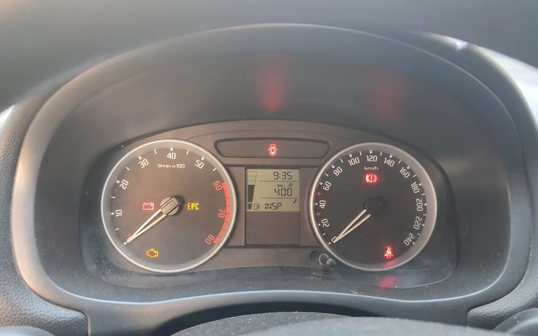 Zbliżenie na deskę rozdzielczą samochodu. Widoczne są dwa okrągłe wskaźniki: po lewej obrotomierz z zaznaczonym czerwonym polem od 6000 obr./min, po prawej prędkościomierz z maksymalną wartością 240 km/h. Pomiędzy nimi znajduje się cyfrowy wyświetlacz pokazujący godzinę 9:35 oraz przebieg 400 km. Na panelu świecą się czerwone kontrolki: akumulatora, układu hamulcowego oraz pasów bezpieczeństwa, a także żółta kontrolka silnika i napis EPC.