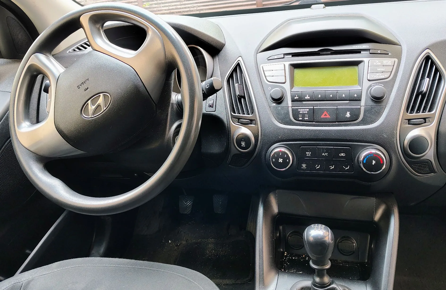 Wnętrze samochodu marki Hyundai, widok na deskę rozdzielczą i kierownicę. Po lewej stronie znajduje się czarna kierownica z logo Hyundai na środku. W centrum deski rozdzielczej widoczny jest system audio z wyświetlaczem, przyciski sterujące oraz panel klimatyzacji z pokrętłami. Poniżej znajduje się konsola środkowa z lewarkiem zmiany biegów. Po obu stronach radia znajdują się pionowe kratki nawiewów.