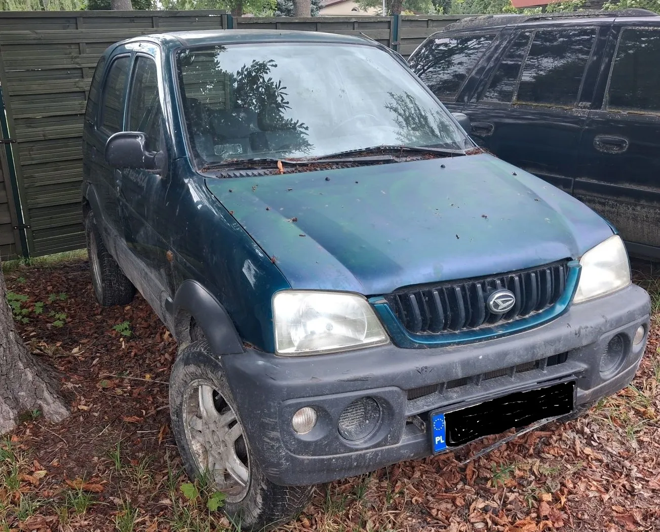 Ciemnozielony samochód osobowy typu SUV marki Daihatsu, zaparkowany na trawie i suchych liściach obok ciemnego ogrodzenia i innego, czarnego samochodu. Pojazd jest zakurzony, z widocznymi zabrudzeniami na masce. Tablica rejestracyjna jest zakryta czarnym polem, widoczny jest jedynie niebieski pasek z oznaczeniem PL.