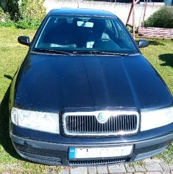 Widok z przodu na czarny samochód osobowy marki Škoda zaparkowany na trawie i kostce brukowej. Widoczna jest maska, przednie reflektory, charakterystyczny grill z logo marki oraz tablica rejestracyjna.