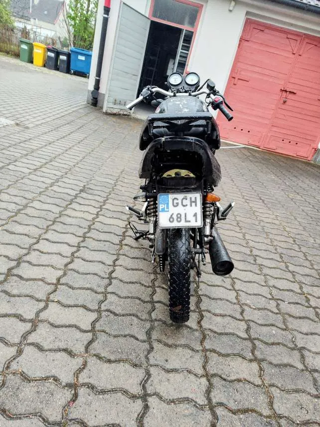 Zdjęcie przedstawia czarny motocykl widziany od tyłu, zaparkowany na brukowanej powierzchni. Na motocyklu widoczna jest biała tablica rejestracyjna z napisem "PL GCH 68L1". W tle znajduje się budynek z czerwonymi drzwiami garażowymi, otwartymi szarymi drzwiami oraz kolorowymi pojemnikami na śmieci (zielony, żółty, czarny, niebieski). Motocykl posiada dwa okrągłe reflektory, czarną siedzisko i brudną oponę tylną. Tło obejmuje również fragmenty domów i drzew.