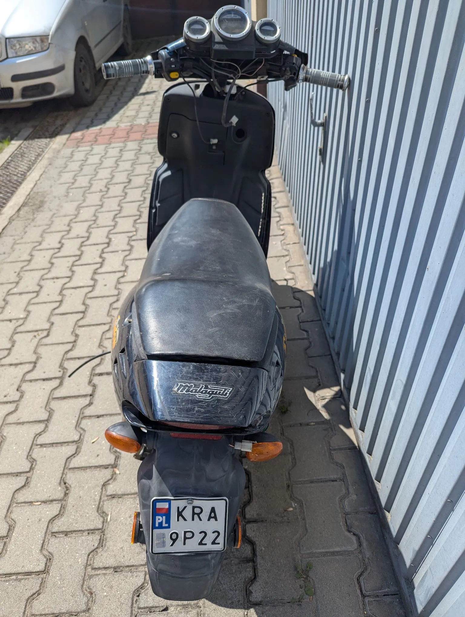 Czarny skuter marki Malaguti zaparkowany na kostce brukowej obok szarej, falistej ściany. Widok od tyłu. Skuter posiada tablicę rejestracyjną o numerze KRA 9P22 z symbolem PL. W tle widoczny jest fragment srebrnego samochodu.