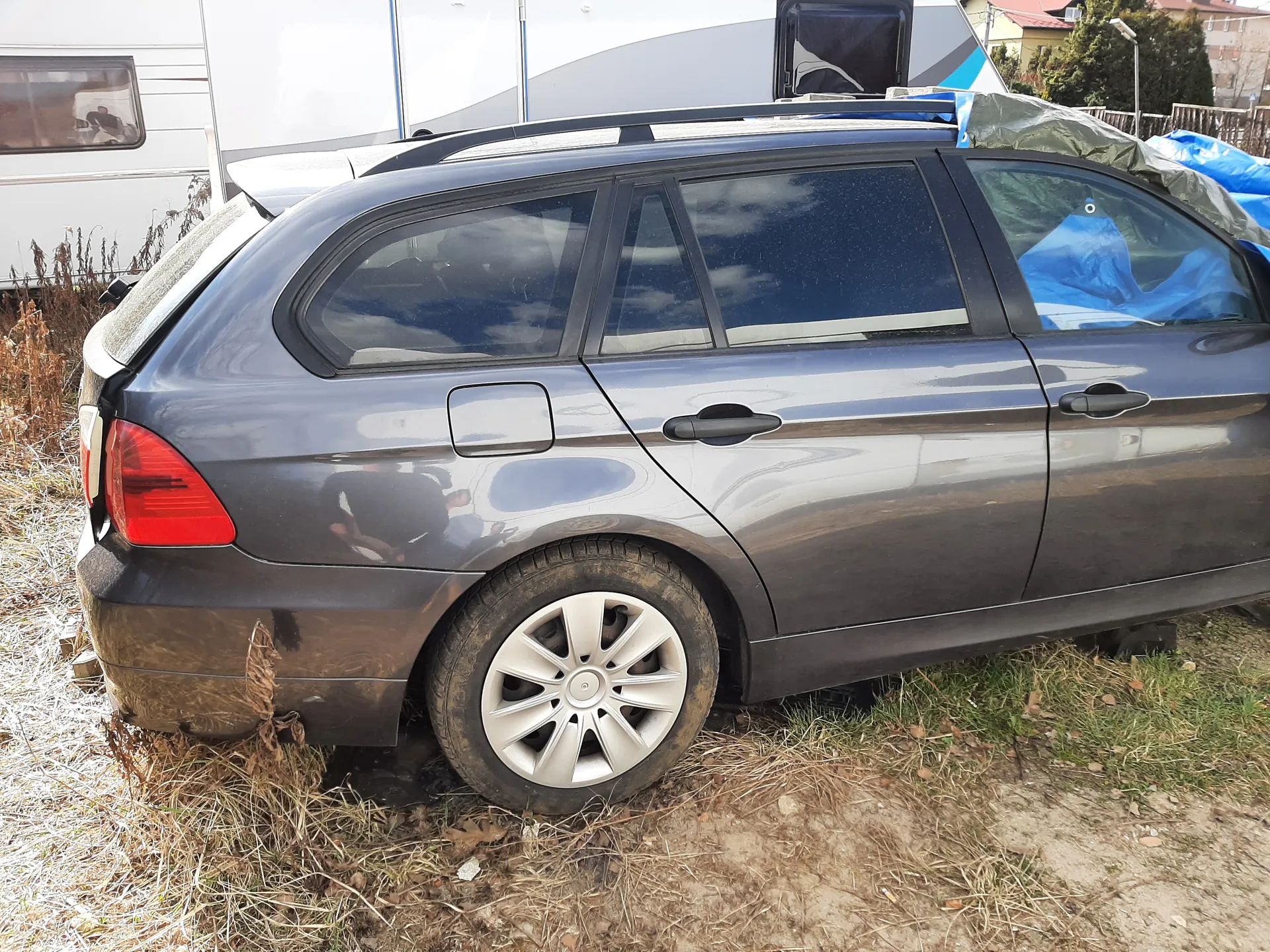 Ciemnoszary samochód typu kombi zaparkowany na trawie i suchych zaroślach. Widoczne są brudne boki pojazdu, czerwone światło tylnego reflektora, srebrne felgi oraz ciemne szyby. W tle biały przyczepa z oknem, niebieskie i zielone folie oraz sucha roślinność. Na ziemi sucha trawa i brudna powierzchnia.