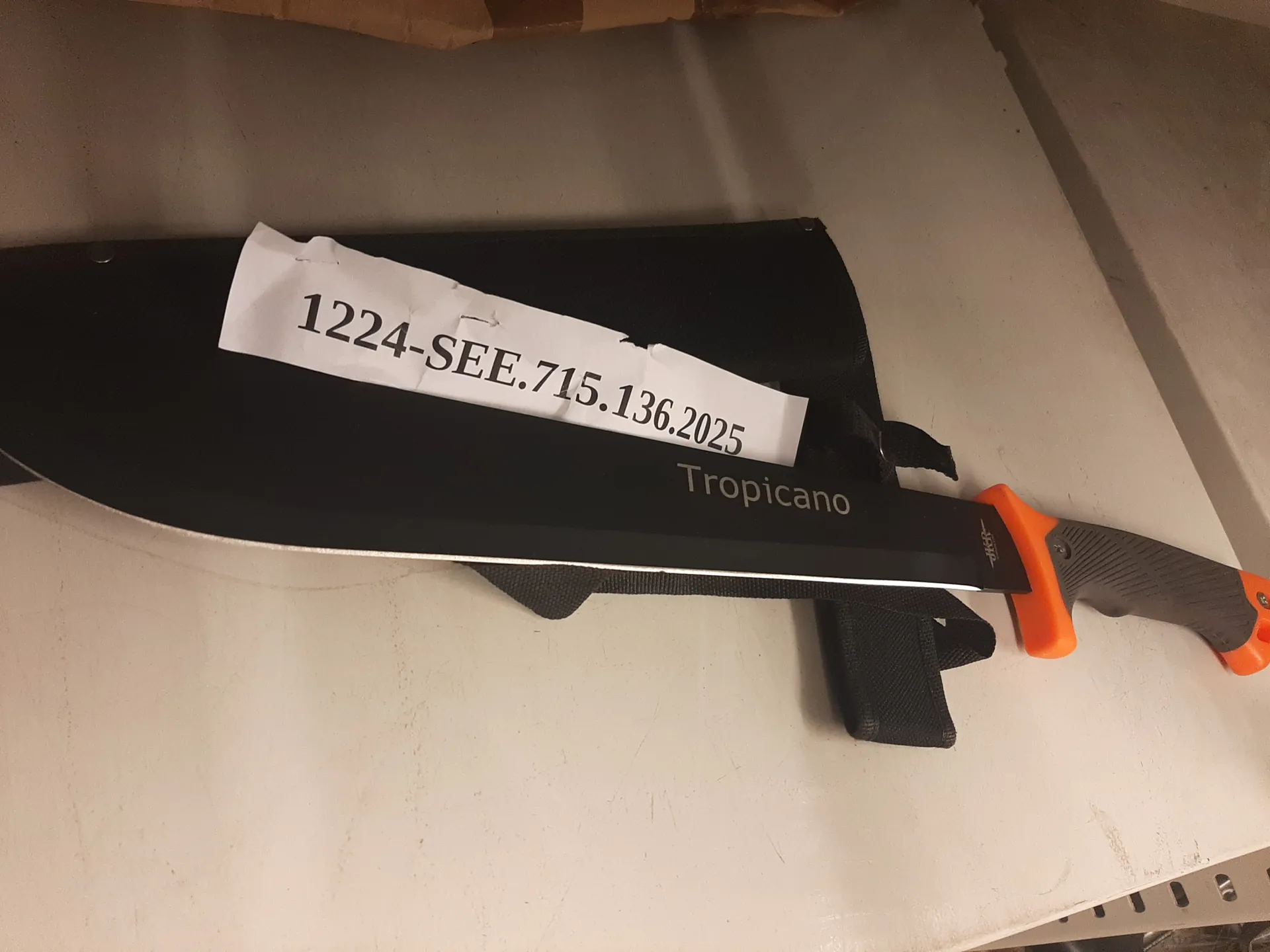 Czarny nóż typu machete z pomarańczowo-czarną rękojeścią leżący na czarnym pochwie. Na ostrzu widnieje napis 'Tropicano'. Na nożu umocowana jest biała taśma z czarnym tekstem '1224-SEE.715.136.2025'. Tło stanowi jasna powierzchnia, prawdopodobnie stół.