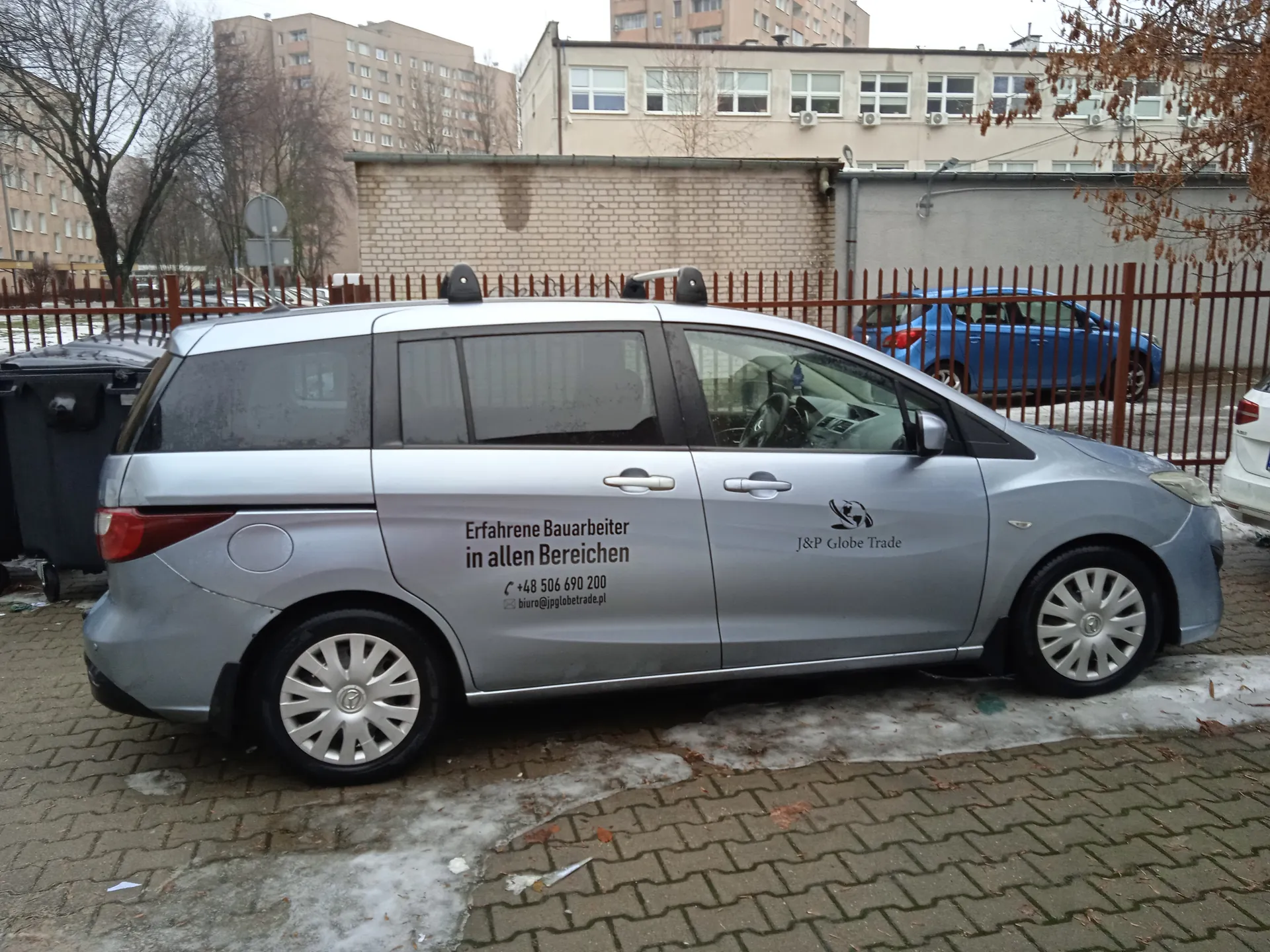 Srebrny samochód osobowy typu minivan zaparkowany na brukowanej powierzchni z resztkami śniegu. Na boku pojazdu widoczny niemiecki tekst: 'Erfahrene Bauarbeiter in allen Bereichen', numer telefonu '+48 506 690 200', adres e-mail 'biuro@jpglobetrade.pl' oraz logo z napisem 'J&P Globe Trade'. W tle: ceglana ściana, metalowe ogrodzenie, niebieski i biały samochód, wielopiętrowe budynki mieszkalne oraz drzewa bez liści. Na ziemi widoczne są plamy śniegu i opadłe liście.