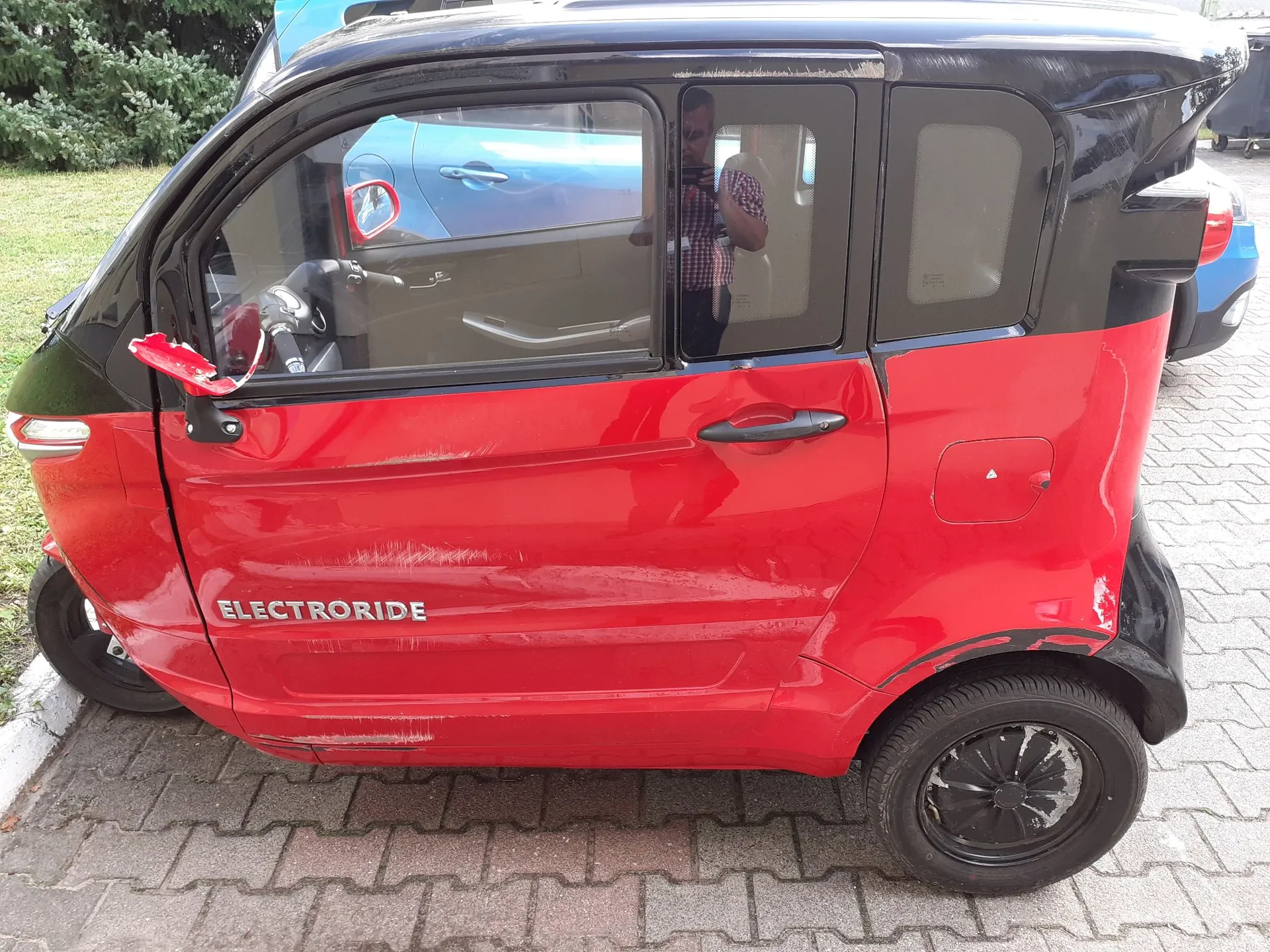 Profil czerwono-czarnego małego samochodu elektrycznego marki Electroride. Pojazd ma czarny dach i górną część nadwozia oraz czerwoną dolną część z napisem 'ELECTRORIDE' na drzwiach. Samochód stoi na kostce brukowej, widoczne są uszkodzenia karoserii w postaci zarysowań na czerwonym lakierze oraz urwane lusterko boczne po stronie kierowcy.