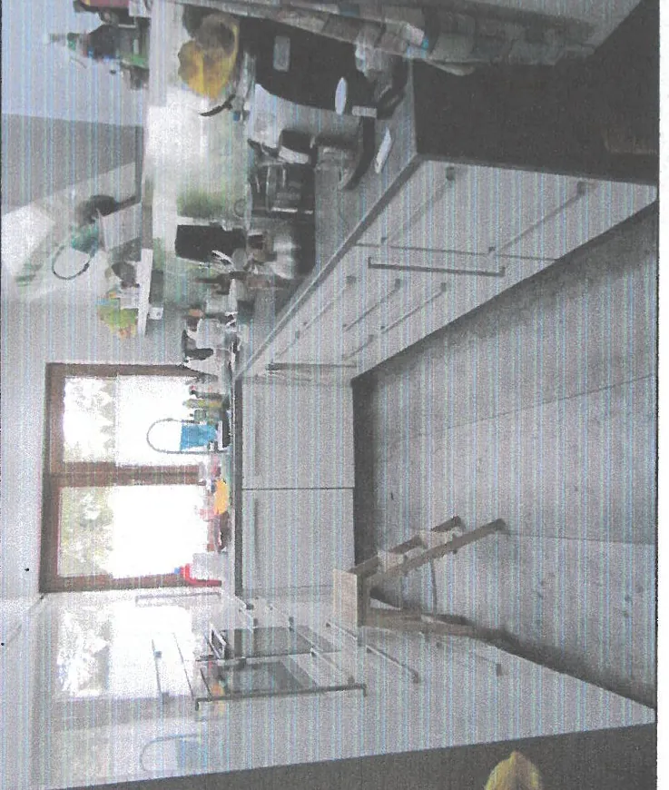 Kuchnia z białymi szafkami, czarną blatem i dużym oknem w tle. Na blacie znajdują się różne przedmioty: czajnik, ekspres do kawy, pojemniki i naczynia. W lewym dolnym rogu stoi drewniana drabinka. Podłoga wyłożona dużymi płytkami. Po lewej stronie widoczna lodówka. Obraz posiada poziome linie przypominające efekt skanowania.