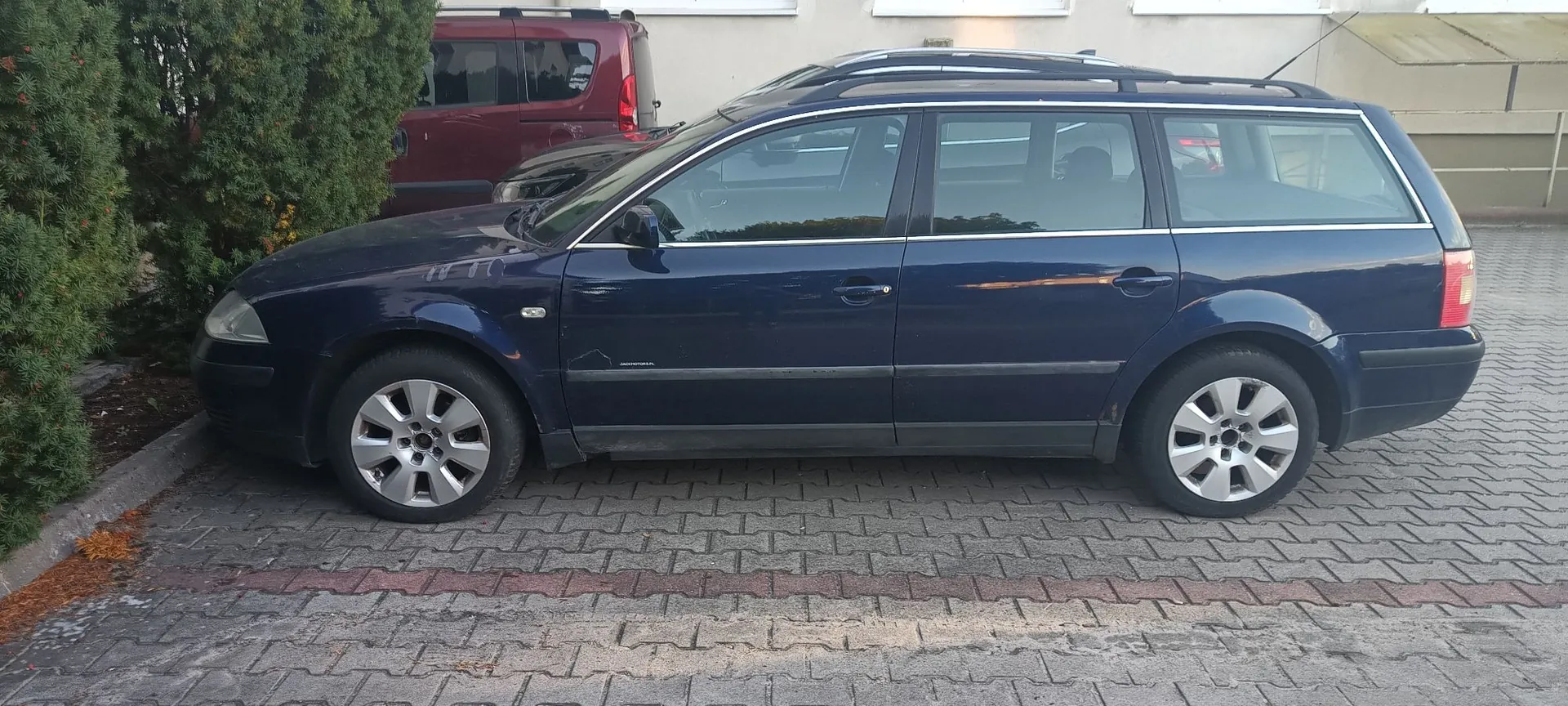 Granatowy samochód osobowy typu kombi zaparkowany bokiem na kostce brukowej. W tle widoczny jest tył czerwonego vana oraz zielone krzewy po lewej stronie.