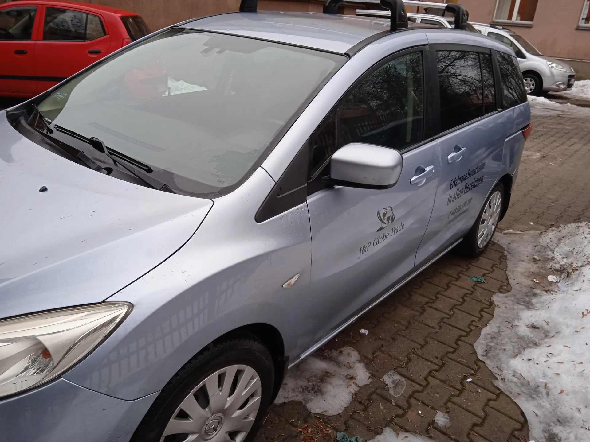 Srebrny samochód typu minivan zaparkowany na brukowanej powierzchni z resztkami śniegu. Na boku pojazdu widoczny napis: "J&P Globe Trade" oraz niemiecki tekst: "Erfahrene Bauarbeiter in allen Bereichen". W tle czerwony i srebrny samochód, brązowa ściana budynku z oknami. Na przedniej szybie refleksje, a na ziemi rozproszone liście i śnieg.