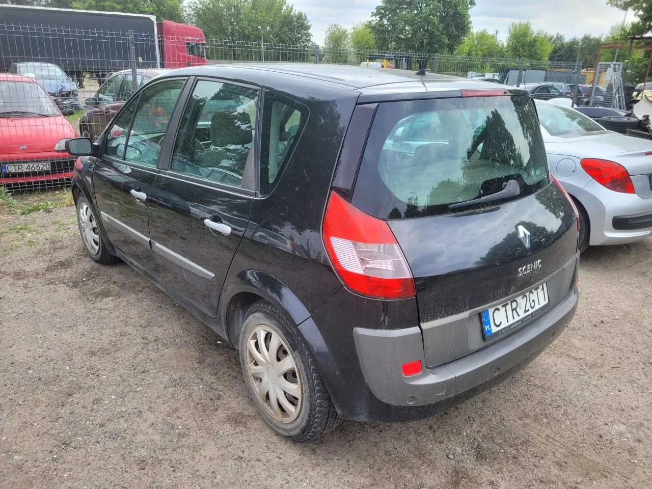 Czarny samochód Renault Scenic z tablicą rejestracyjną CTR 2G11 (PL) zaparkowany na żwirowym placu. W tle widoczne są inne pojazdy: czerwony samochód z tablicą CTR 46620, srebrny samochód oraz ciężarówka. Obok znajduje się ogrodzenie, drzewa i chmurne niebo. Tekst "Scenic" widoczny na tylnej części pojazdu.