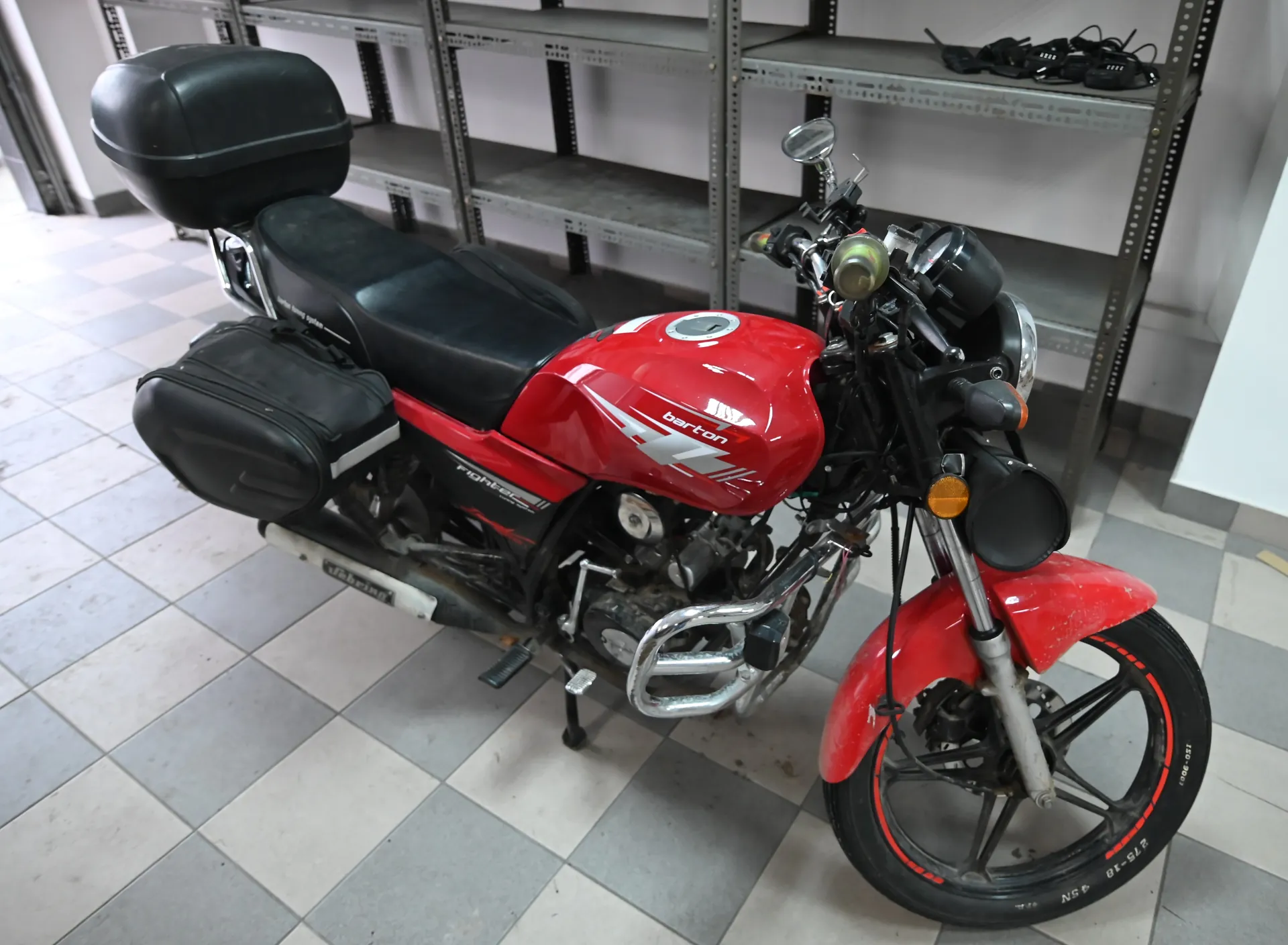 Czerwony motocykl Barton Fighter z czarnym siedzeniem, bagażnikiem i boczną torbą. Widoczne napisy "Barton" na zbiorniku paliwa i "Fighter" na ramie. Stoi na szaro-białej mozaikowej podłodze, w tle metalowe półki z przedmiotami.